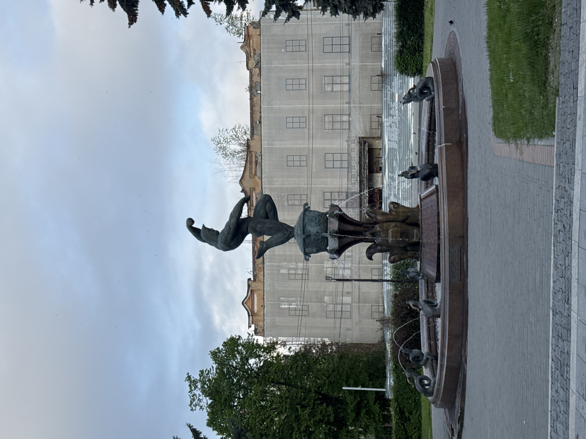 The image captures a view of the Mikhail Avakyants Fountain in Akhieli, Georgia. The fountain features a bronze sculpture of two intertwined figures, one seemingly airborne and the other grounded. The fountain itself has multiple levels and spouts water from the mouths of animal figures into a circular basin. Behind the fountain is a building undergoing renovation, covered in netting with outlines of windows. To the sides are paved walkways and grass areas, with trees and bushes adding greenery to the scene. The sky is overcast, suggesting a cloudy day.