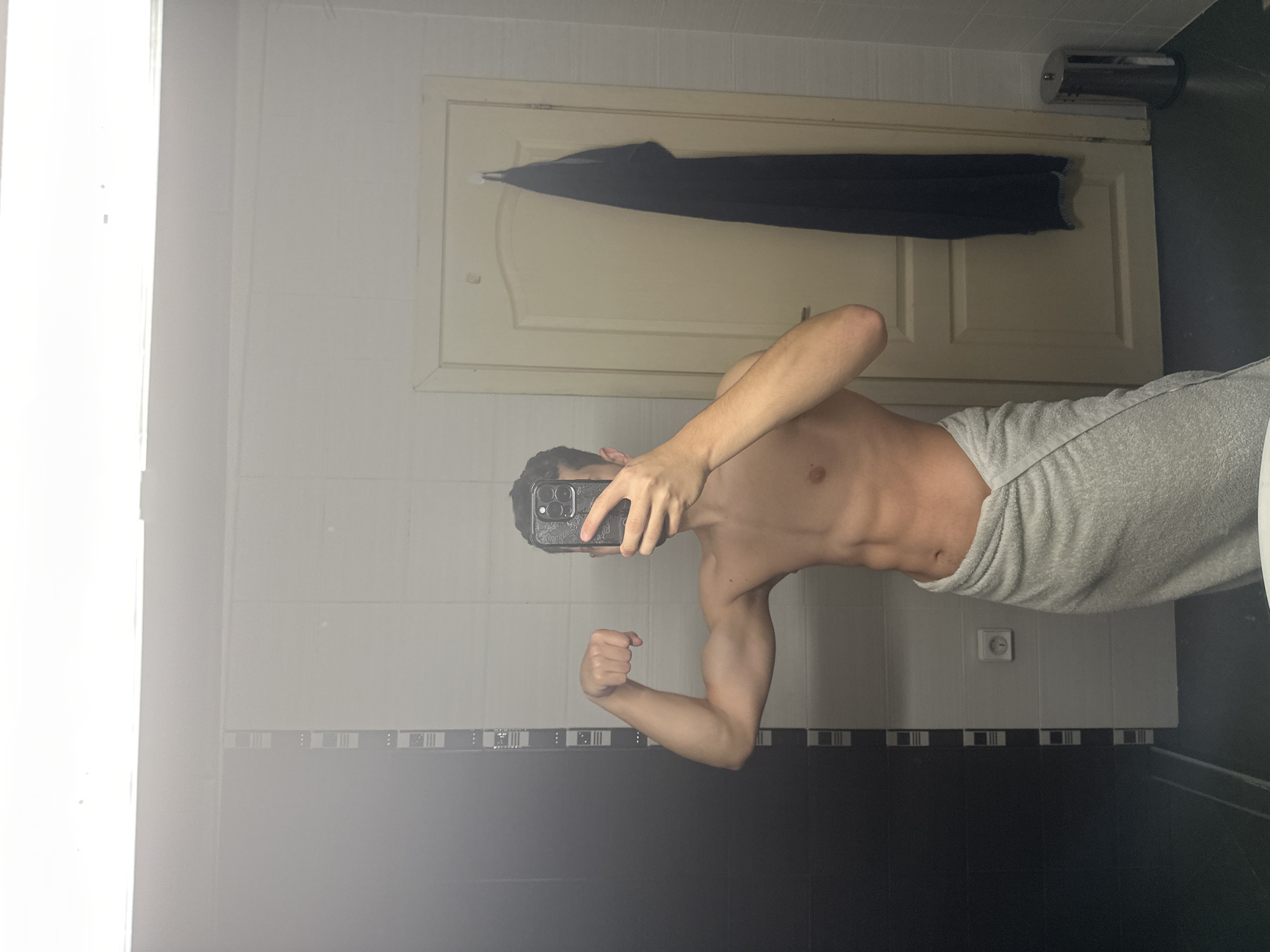 A person, appearing to be a young male, takes a shirtless selfie in a mirror within a bathroom located in Unknown, Unknown. He is wrapped in a light grey towel from the waist down, obscuring his lower body. His left hand holds a dark-colored smartphone, primarily an iPhone due to the visible camera module, in front of his face, while his right arm is bent, flexing his bicep. He has dark hair and a lean physique with visible abdominal muscles. The bathroom features a dark grey tiled floor. The walls reflected in the mirror exhibit a pattern of alternating black and white vertical rectangular tiles on the left. To the right, a light-colored wooden door with a white frame is visible, from which a dark blue towel hangs on a white hook. A white electrical outlet is mounted on the wall to the left of the door. A silver cylindrical trash can with a lid stands on the floor to the right of the person, near the door. A bright, elongated light fixture is reflected horizontally across the top of the mirror.