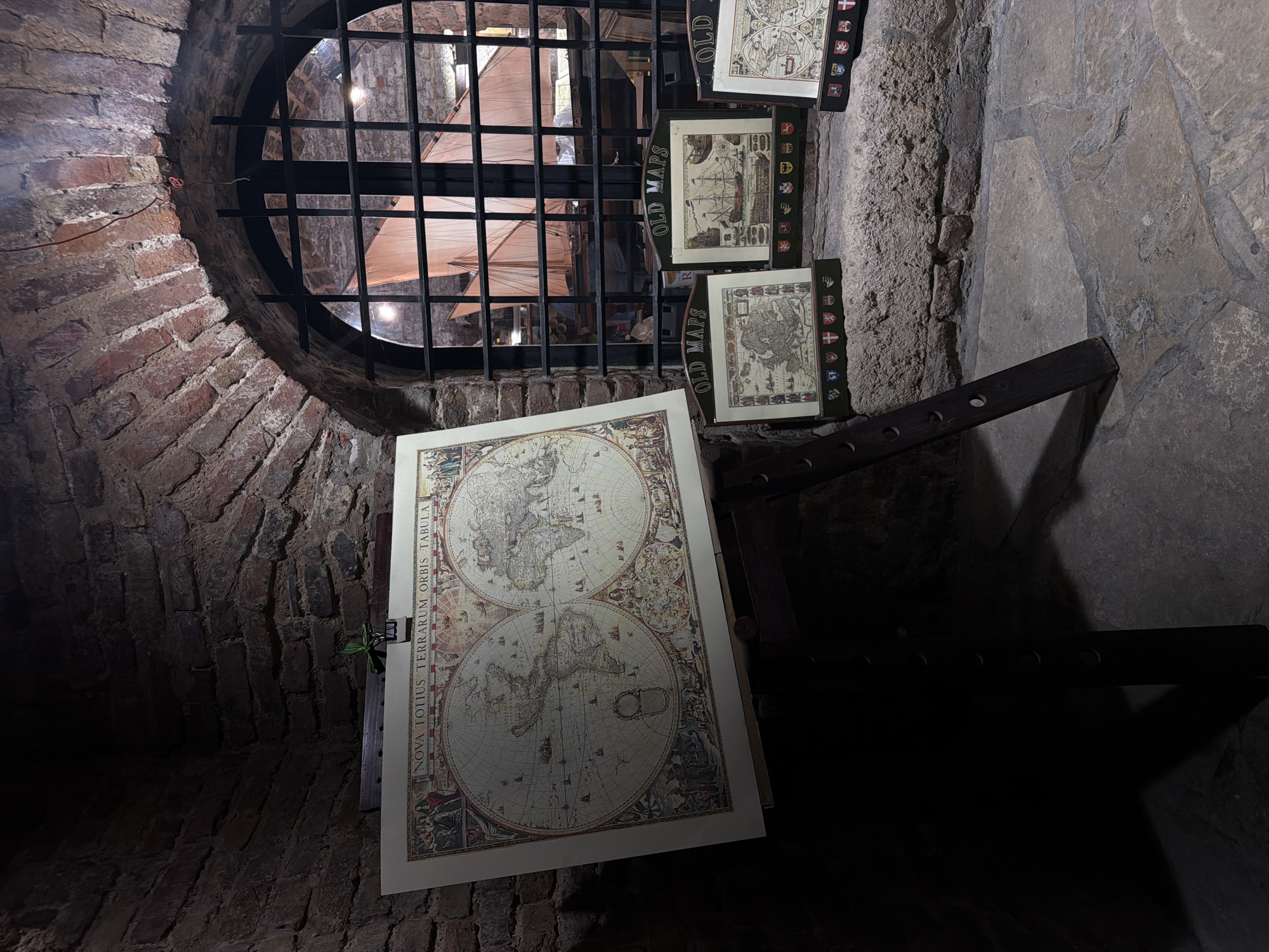 The image shows a dimly lit interior of a building, featuring stone walls constructed with brick and stone. A barred, round window is located on the left side, revealing an exterior view of a large sail-like structure, possibly belonging to a ship or architectural element. In the foreground, a large map depicting a world map is leaning against the wall. Adjacent to this, three smaller maps or framed images are displayed on what appears to be a rack or support structure made of dark wood. The location is Belgrade, Serbia, as indicated by the map details.