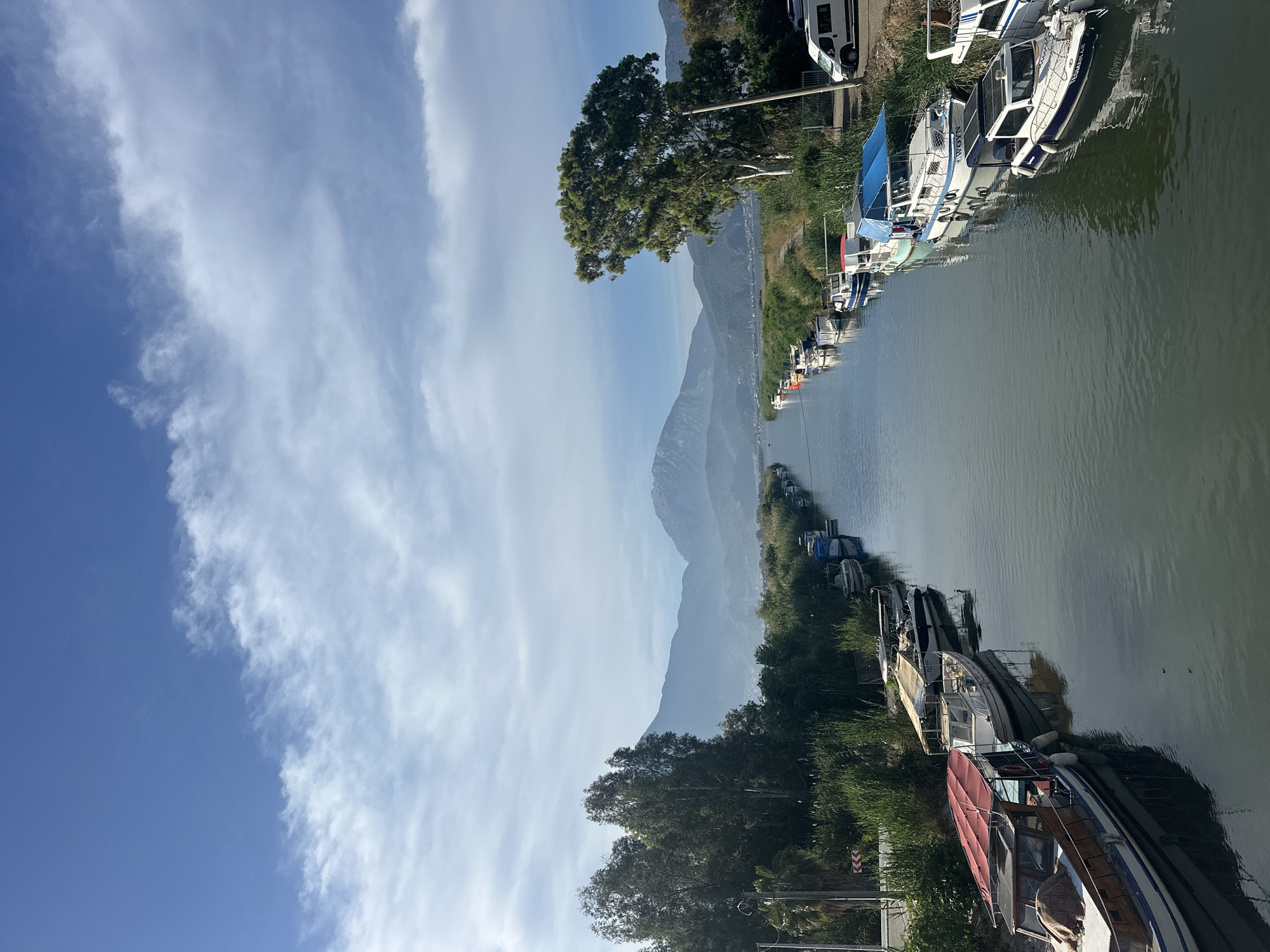 The image shows a waterway lined with boats in Fethiye, Turkey. The waterway is positioned between two banks of greenery. On one bank, boats are moored close together, while on the opposite bank, vegetation leads to a mountain range in the distance. The sky is partly cloudy, transitioning from blue to a lighter hue near the mountains. Along the shoreline, boats of varying sizes and designs are visible. Some appear well-maintained, while others show signs of wear. The water's surface reflects the sky and surrounding landscape.