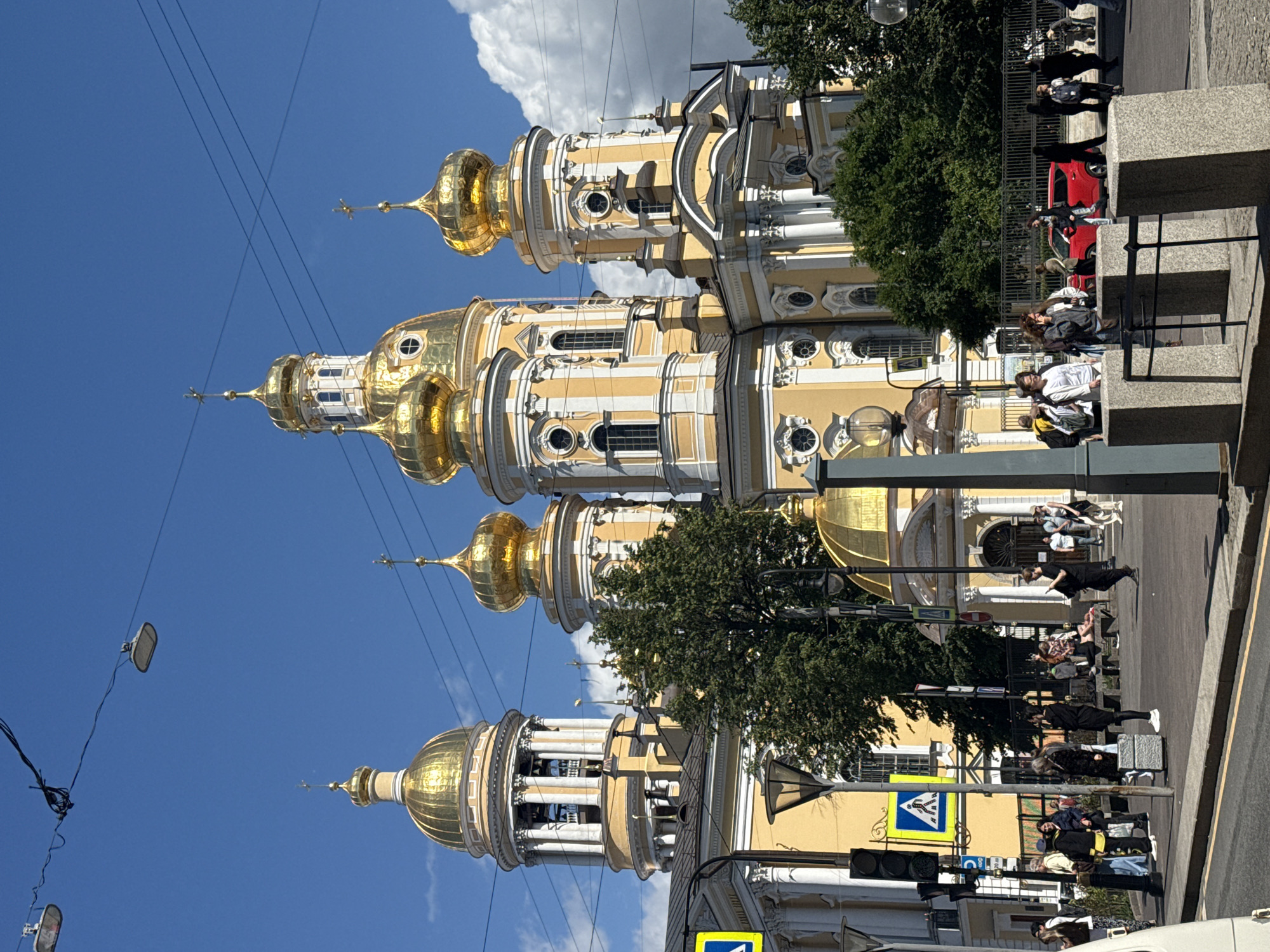The image depicts the Church of the Resurrection of Christ, also known as the Church of the Savior on Spilled Blood, located in Saint Petersburg, Russia. The church is characterized by its ornate, onion-shaped golden domes and its predominantly yellow facade, adorned with white architectural details. The scene captures the front of the church on a sunny day with a bright blue sky and scattered clouds.
In the foreground, a street and sidewalk area is visible with several people present. Some individuals are walking, while others are standing and conversing. Traffic lights and pedestrian crossing signs are also present. A bright red car is parked on the street.
The church's distinctive architecture dominates the background, with multiple golden domes and spires reaching towards the sky. Green trees are interspersed around the church and along the street. Overhead, power lines and what appear to be streetlights are strung across the sky. The overall impression is a bustling urban environment with a prominent historical and religious landmark.