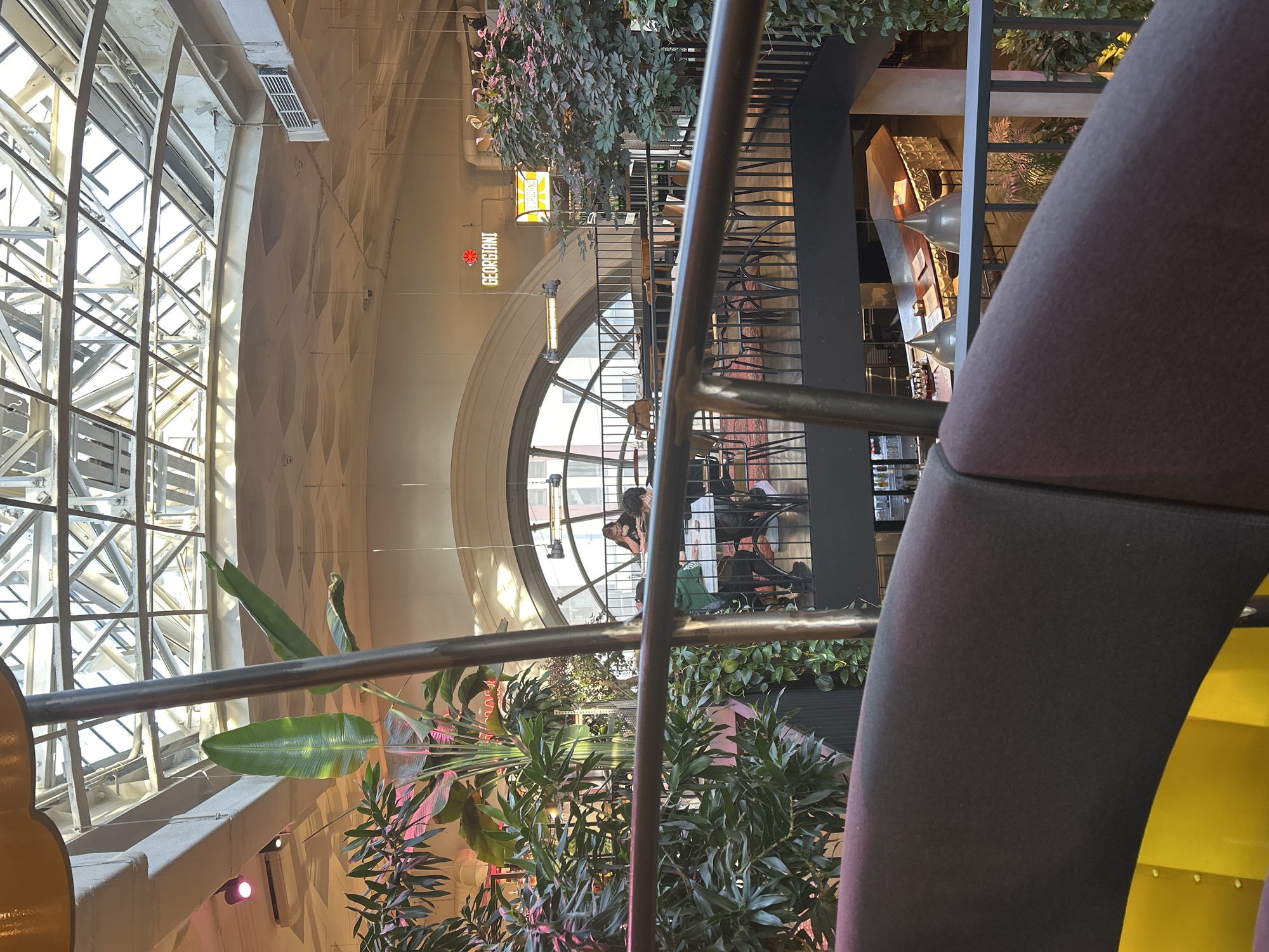 The image depicts an interior view of a multi-level establishment, possibly a restaurant or a public space, with a prominent glass ceiling supported by a metal lattice structure. Large, lush green plants are strategically placed throughout the levels, adding to the ambiance.
On the upper level, several individuals are seated at tables, engaged in conversation or dining. One person is visibly looking at a device. Further in the background, a sign reads "GEORGIANI" in neon letters, accompanied by a smaller sign with yellow lettering.
The middle level features a railing overlooking a lower section of the establishment. More tables and chairs are visible, along with a red patterned rug.
The lower level shows a bar area with a curved wooden counter, pendant lamps, and bar stools. Bottles are arranged on shelves behind the bar.
The foreground of the image is dominated by the dark fabric of seating and a bright yellow surface. The overall impression is a modern, airy space with abundant greenery and a lively atmosphere. The specific city and country are not identifiable from the image.