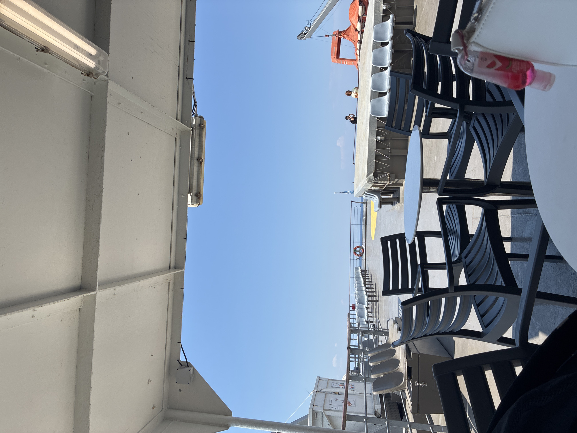 The image shows an outdoor seating area on the deck of a ferry, likely en route to or from the Municipal Unit of Kastro - Kyllini, Greece. The foreground features several dark plastic chairs around white tables, some of which are occupied by bags and a bottle of hand sanitizer. In the middle ground, rows of empty, light-colored plastic seats are arranged facing the sea. Beyond the seating area, a life raft and a crane are visible on the ferry's deck. Two individuals, a man and a woman, are standing on a raised platform in the distance, observing the surroundings. The background is dominated by a clear blue sky and the calm sea. The deck is covered by a white canopy, providing shade. The overall scene suggests a sunny day at sea with passengers enjoying the view.