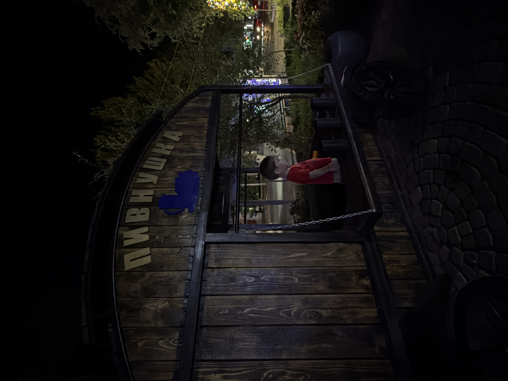 The image captures a night scene in the city of Chirchik, Uzbekistan. In the foreground, a young boy wearing a red t-shirt stands inside a wooden structure resembling a small kiosk or cart. The structure has wooden planking and a curved roof with the word "ПИВНУШКА" inscribed in yellow letters. A blue silhouette of a beer mug is visible above the text. The boy is facing to the left, looking down. The structure has a window opening where the boy is standing, and a chain hangs from the top of the opening.
The ground in front of the structure is paved with cobblestones arranged in a circular pattern. In the background, trees are visible, some illuminated by what appears to be decorative lighting, including a tree decorated with yellow lights. Further in the background, city lights and the headlights of a few cars on a street can be seen, suggesting the location is near a road. The overall lighting suggests it is nighttime, with artificial light sources illuminating the scene.