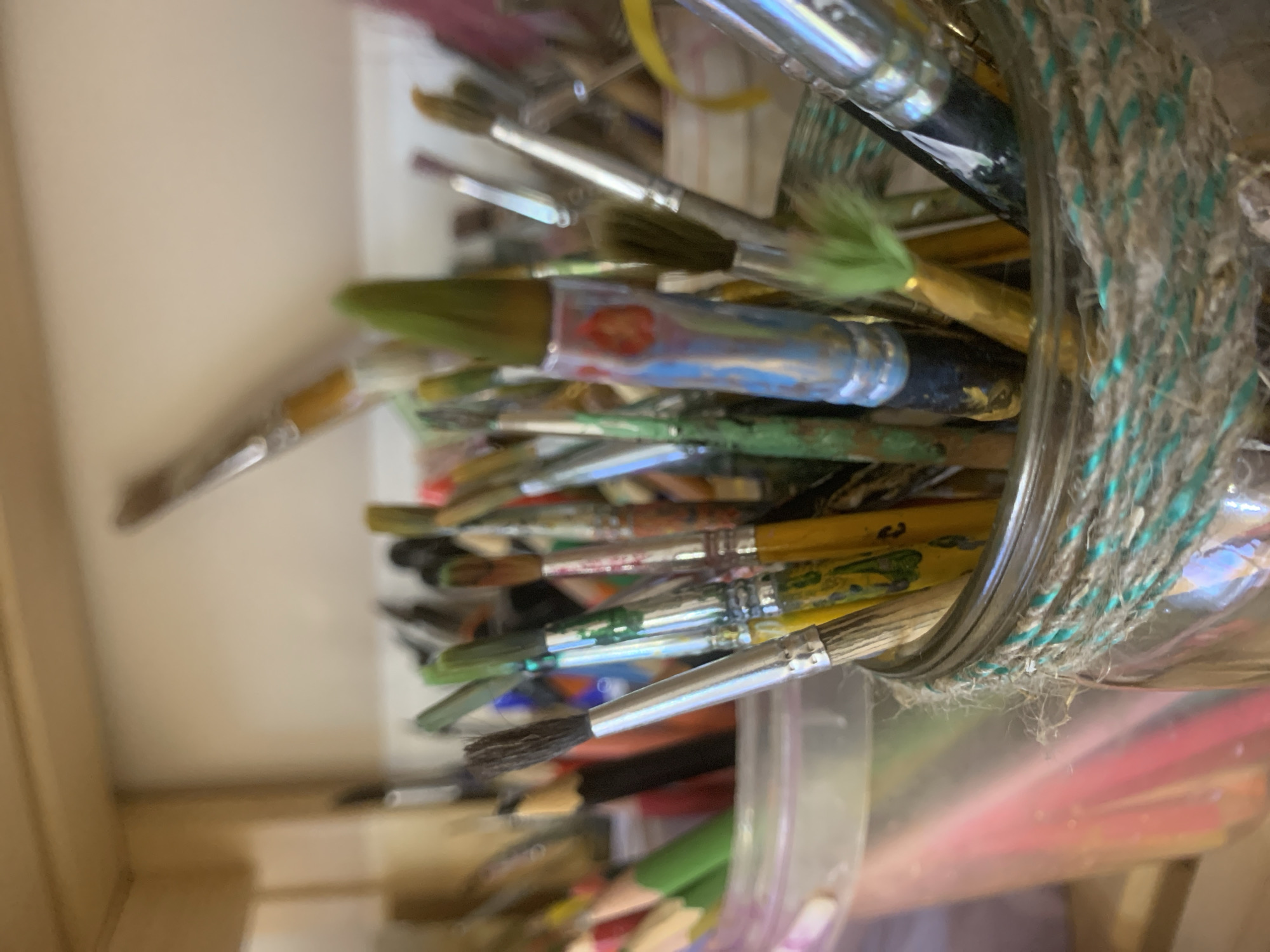 The image shows a jar filled with paintbrushes and colored pencils. The jar is wrapped with a rustic twine that has a blue and white pattern. The paintbrushes are various sizes and show signs of use with paint residue on the bristles and handles. The colored pencils are visible at the bottom of the jar. The scene appears to be indoors. There is no indication that this image is located in the city of Izhevsk, Russia.