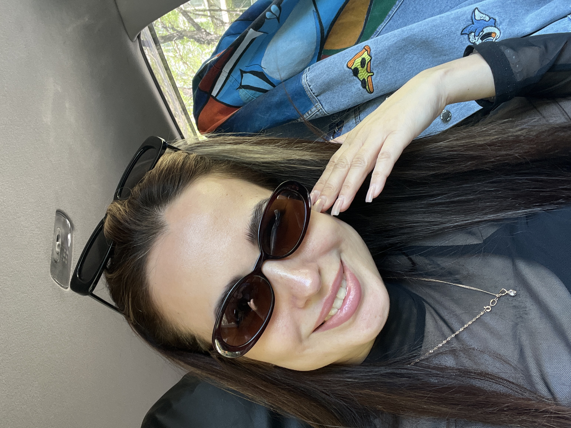 A woman is photographed in what appears to be a car. She is wearing sunglasses and smiling. There's a jacket with patches (a pizza slice and a character) behind her. There's a rearview mirror and the car's interior visible in the background. Location is Unknown, Unknown.
