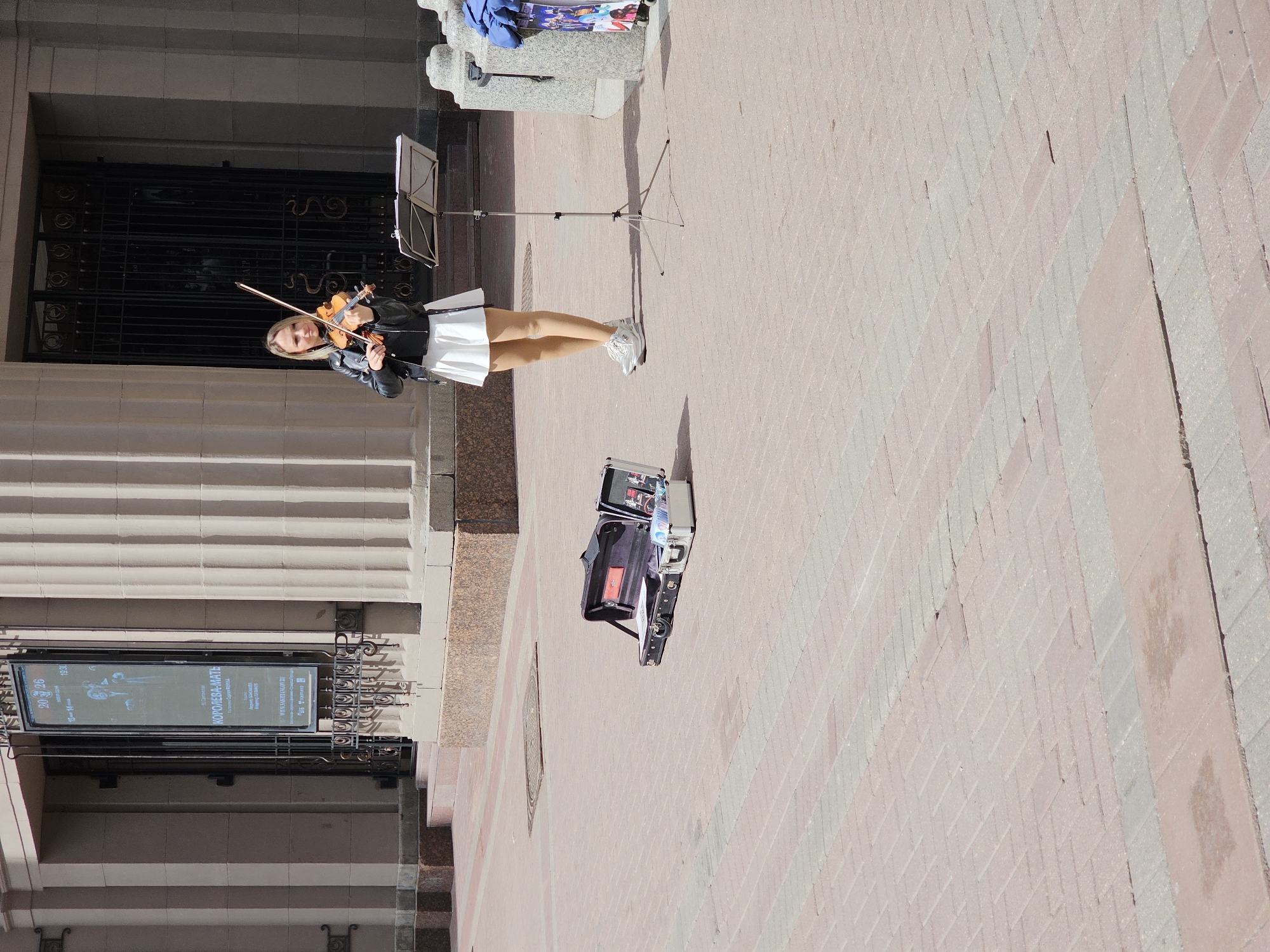 The image shows a street performer playing the violin on a brick-paved sidewalk in Moscow, Russia. She is a blonde woman wearing a black leather jacket and white skirt. She stands in front of a music stand, with an open suitcase containing equipment nearby. Behind her is a building with ornate metalwork and a sign that reads "КОРОЛЕВА-МАТЬ," which roughly translates to "Queen Mother." The sign also displays dates 20 and 26, likely indicating a performance or event schedule. A column and decorative stone structure is also visible, located close to the street performer. The light suggests a sunny day.