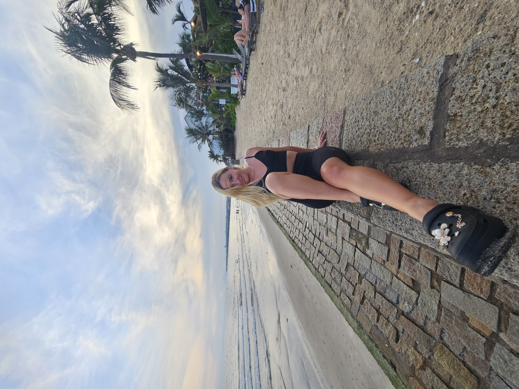A young woman with blonde hair is sitting on a stone embankment at a beach in Phu Thuy Ward, Vietnam. She is wearing a black sports bra and black shorts, and black clogs decorated with charms. The beach has sand and palm trees in the background. There are lounge chairs with people on them further down the beach. The sea is calm with small waves. The sky is cloudy with the sun setting or rising.
