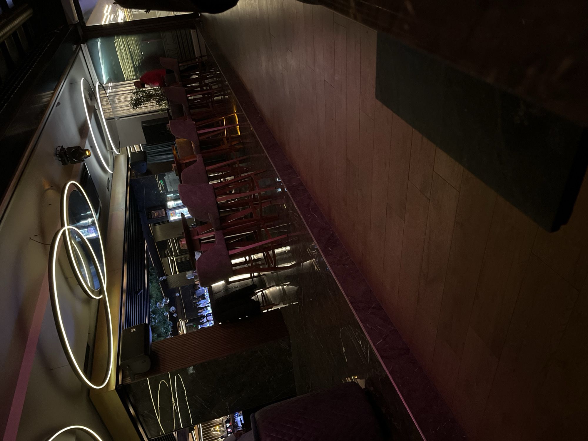 The image depicts the interior of a modern establishment, likely a restaurant or bar. Rows of tables and chairs are aligned along a reflective surface, suggesting a polished floor. There are light fixtures with circular neon lights on a wall. The color palette is dark, with strategic lighting accentuating certain areas. The setting could be in Plovdiv, Bulgaria, but no specific landmarks confirm the location. There are no readily discernible people or actions visible in the image.