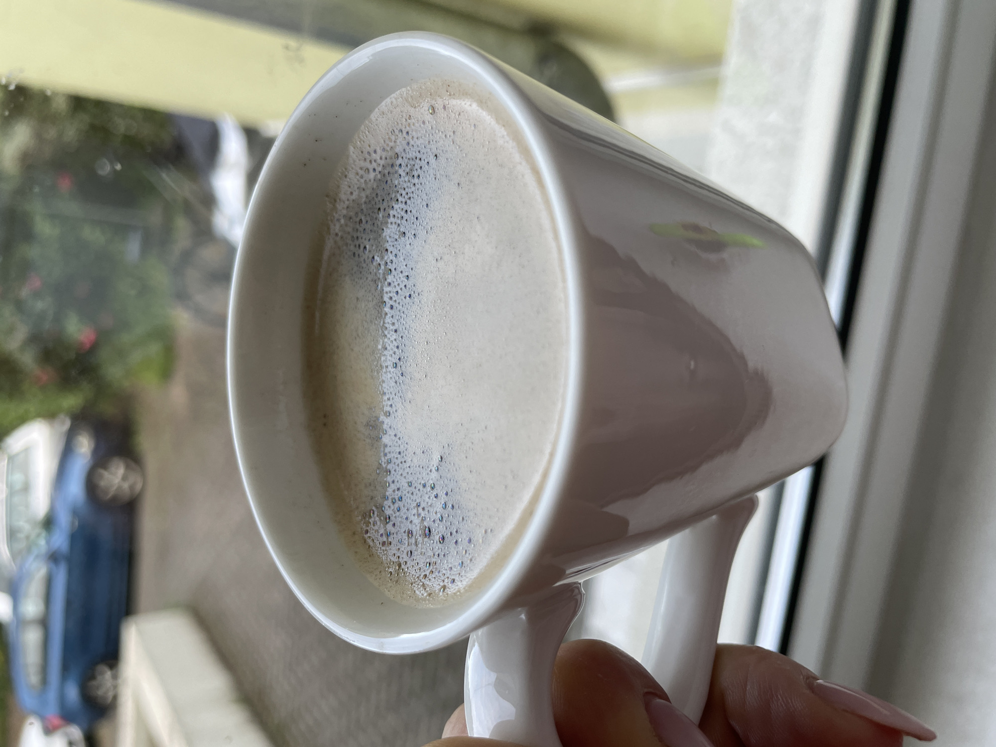 A close-up shot shows a white mug of coffee held by a person with light pink painted fingernails. The mug is filled with a light brown coffee with a frothy top layer. In the background, through a window, a blue car is parked on a cobblestone driveway. Green foliage and a yellow wall are also visible outside. The scene appears to be from a residential area.
