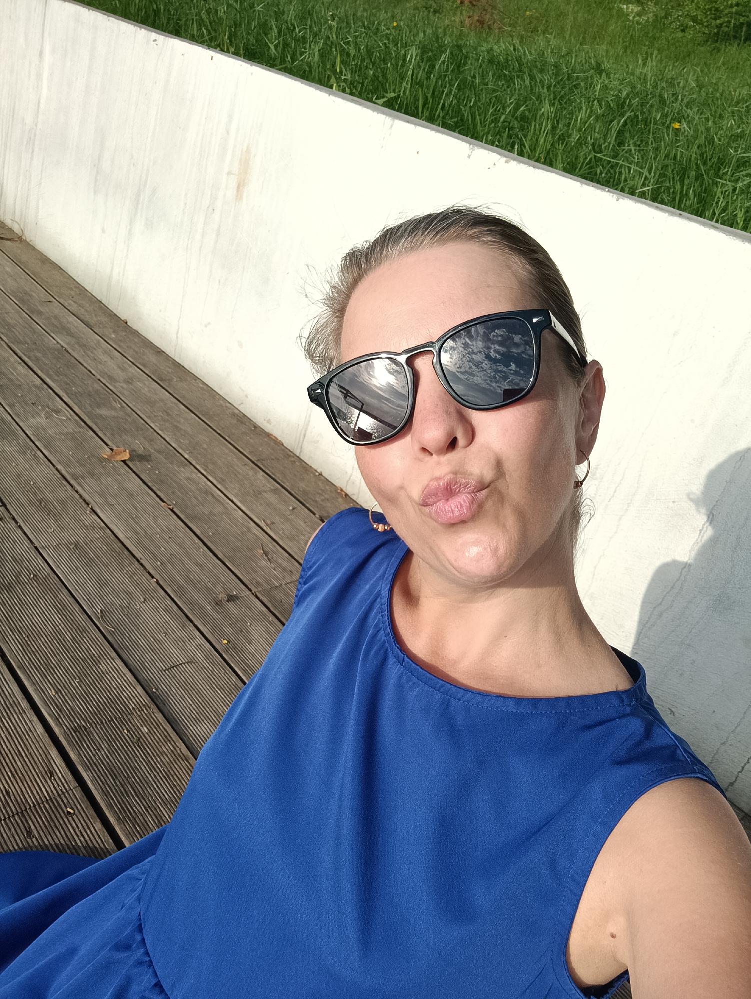 The image depicts a woman wearing sunglasses and a blue dress, sitting on a wooden deck next to a white wall. In the background, there is a grassy area. The woman has her lips pursed as if posing for a photo. The sunglasses reflect the sky with clouds. Based on geolocation clues, the photograph might have been taken in the city of Chaikovsky, Russia, though it is impossible to confirm from the image alone.
