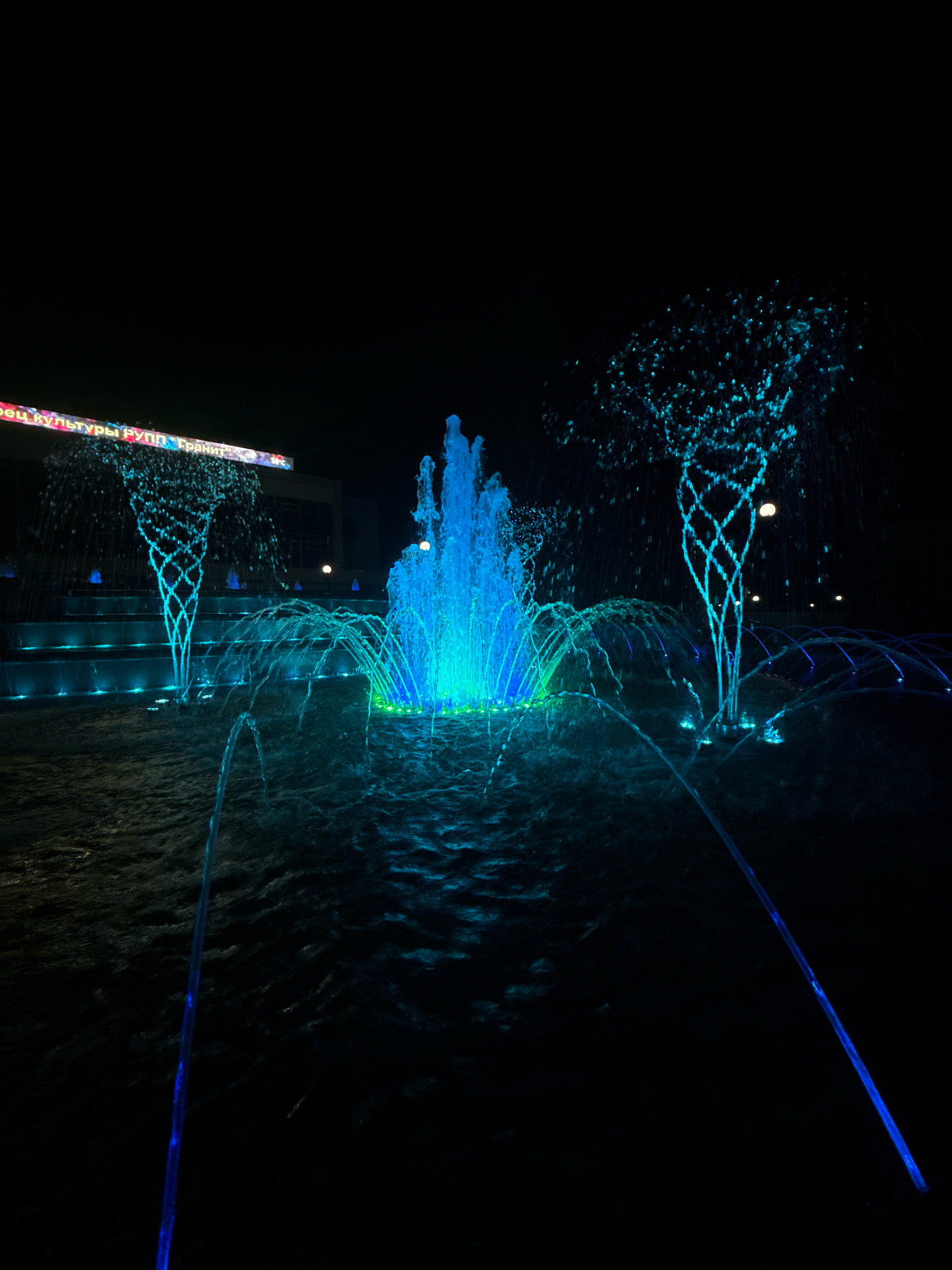 The image depicts a night scene of a fountain in the city of Mikashevichy, Belarus. The fountain is illuminated with blue and green lights, creating a vibrant and dynamic display. Water jets spray upwards, forming various shapes and patterns. In the background, there is a building with a sign that appears to be in Cyrillic script, illuminated with colorful lights. The ground around the fountain is wet, indicating the presence of water. The overall atmosphere is one of a lively public space at night.