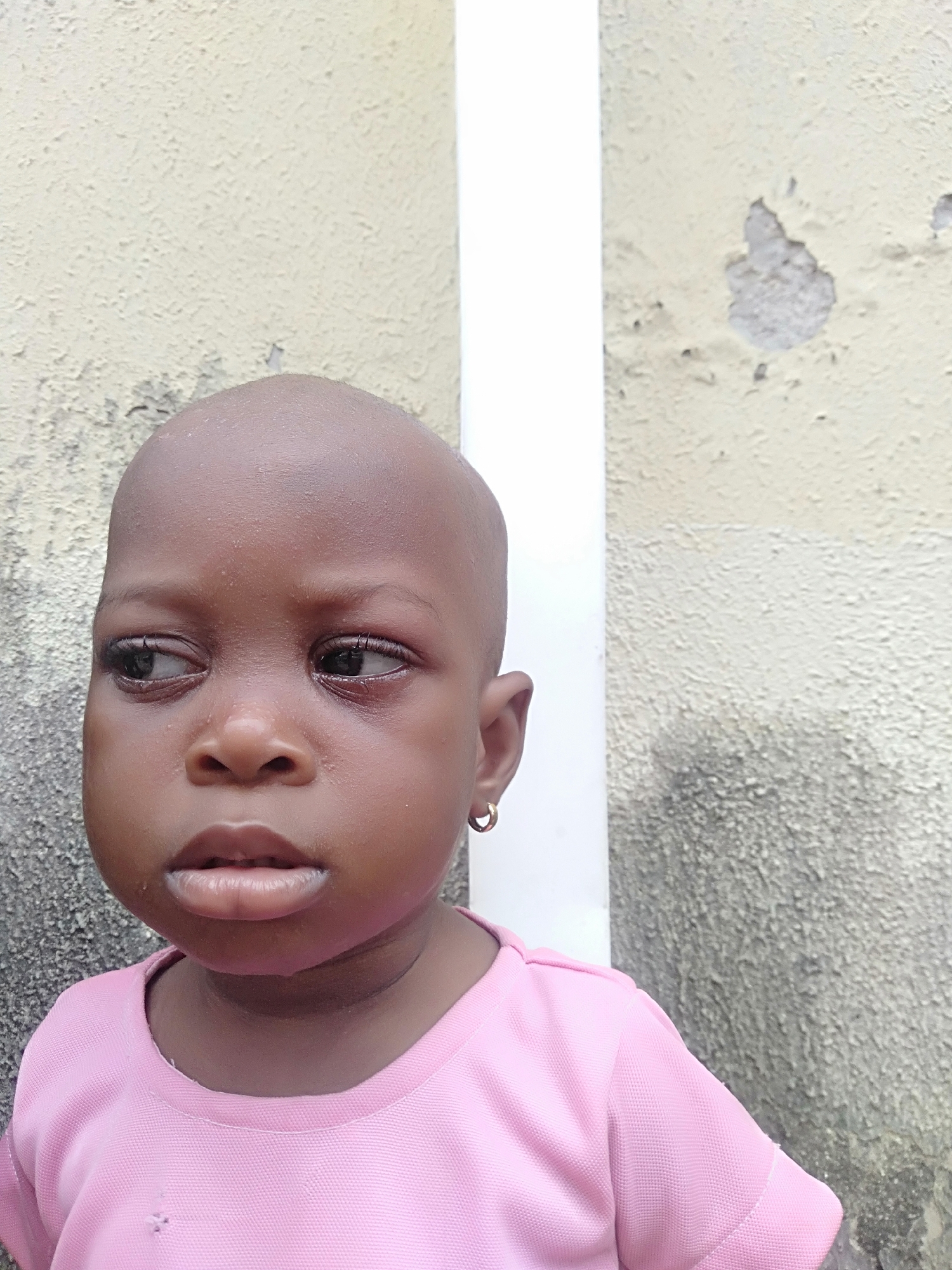 A bald, dark-skinned child wearing a pink shirt and gold hoop earring is positioned in front of a light-colored, textured wall with peeling paint. The child is looking slightly to the left. The location is implied to be Shangisha, Nigeria.