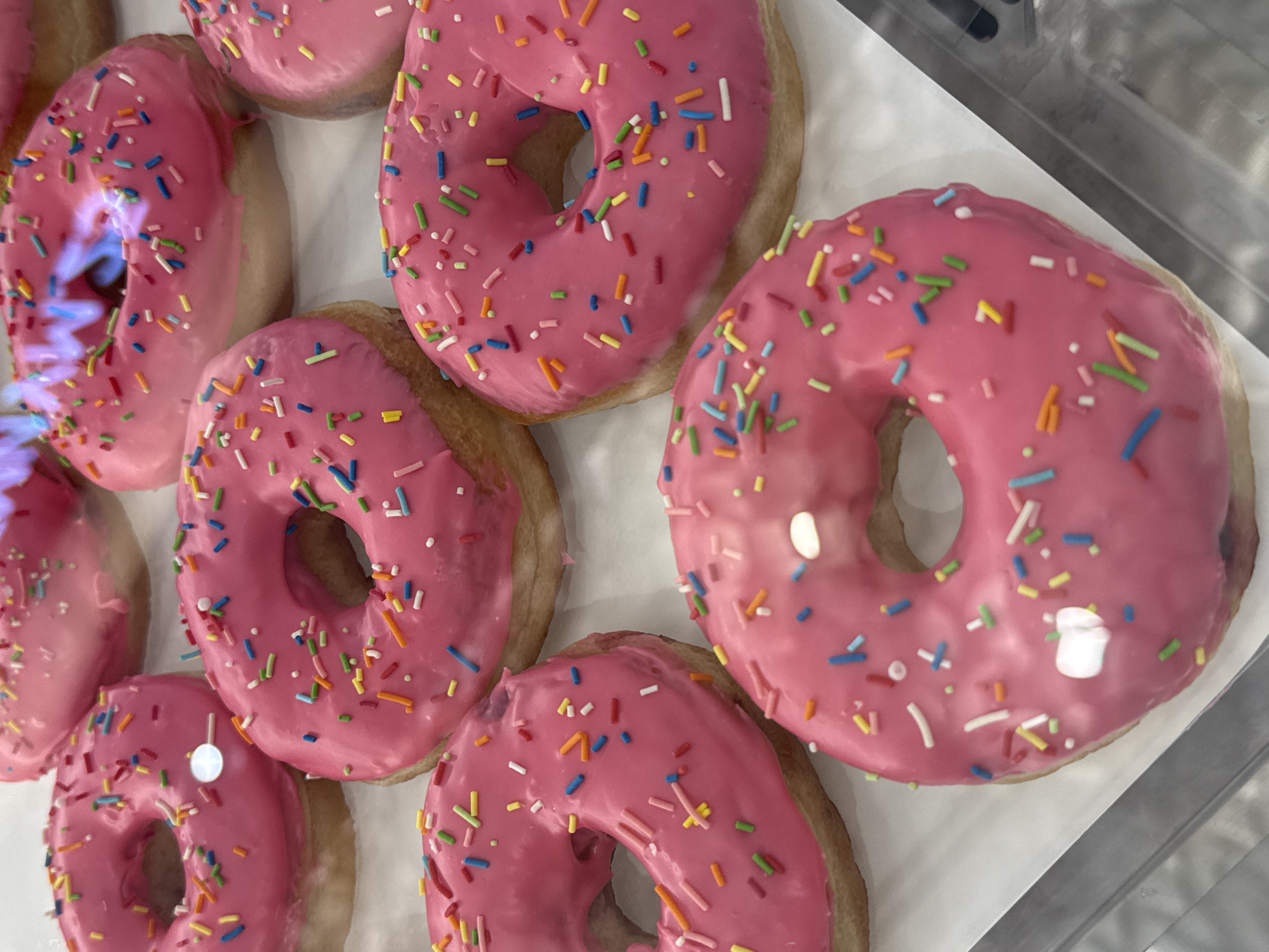 A high-angle, close-up shot reveals a display of seven pink-frosted donuts, each adorned with multicolored sprinkles. The donuts appear to be arranged on a white surface, potentially a tray or display case liner. The lighting is bright, accentuating the pink hue of the frosting and the colorful sprinkles. The reflective glare on the donuts suggests a glass or transparent barrier, potentially part of a display case. The setting is possibly a bakery or cafe, and the location is unknown, making it impossible to confirm if it is in Vladivostok, Russia.