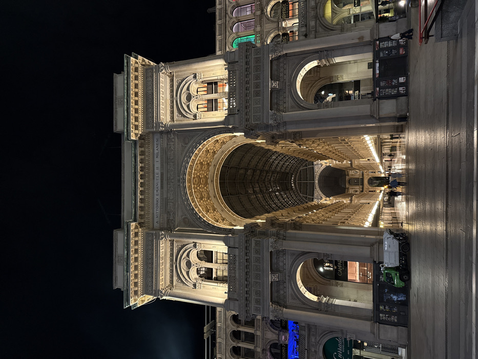 This nighttime, low-angle photograph captures the grand arched entrance to the Galleria Vittorio Emanuele II in Milan, Italy, specifically from the Piazza del Duomo side. The elaborate neoclassical facade, illuminated by warm artificial light, dominates the frame, showcasing ornate architectural details, columns, and a pediment inscribed with "A VITTORIO EMANUELE II. I MILANESI." Beyond the main arch, the long, glass-roofed interior of the shopping gallery is visible, lined with warmly lit storefronts. The ground leading into the gallery is paved and reflects the artificial light.
On the left side of the entrance, a small, green and white utility or waste collection truck is parked. Nearby, a dark kiosk or newsstand is present. Several individuals are visible walking within the brightly lit entrance area of the Galleria, moving away from the viewer. On the far right, another person is visible walking near a similar kiosk. Buildings adjacent to the main entrance feature various illuminated windows and signs, including a blue sign on the left and windows with green and orange lighting on the right. The sky above is dark, indicating a clear night.