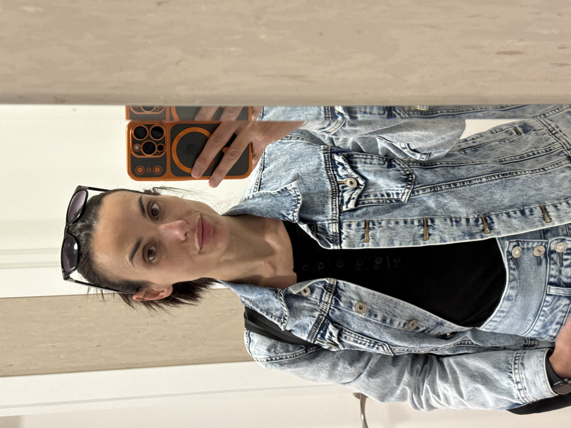 A woman stands in front of a mirror taking a selfie. She is wearing a light blue denim jacket, a black top, and sunglasses perched on her head. She holds an orange and black phone in her left hand to capture the image. The background appears to be a restroom with white or light-colored surfaces. The city of Pleven, Bulgaria cannot be confirmed from the image.