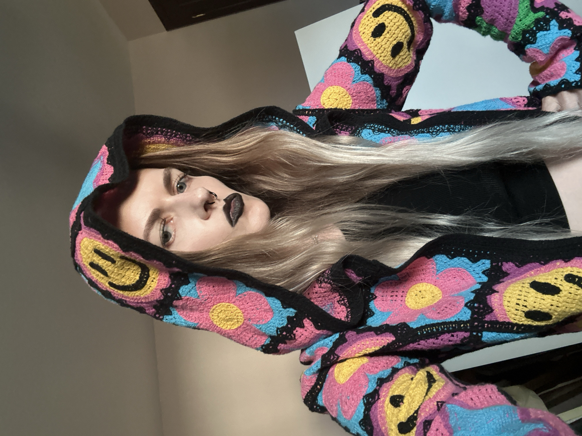 A young adult female with long, light blonde hair, dark lipstick, and a nose ring is pictured from a close-up, tilted angle. She is wearing a black top and a hooded jacket or sweater. The jacket features a colorful crochet pattern of pink flowers, blue accents, yellow smiley faces, and black trim. The background shows a plain wall of light color and a white surface. The location is unknown, it is not possible to tell that this image was taken in Cape Coral.