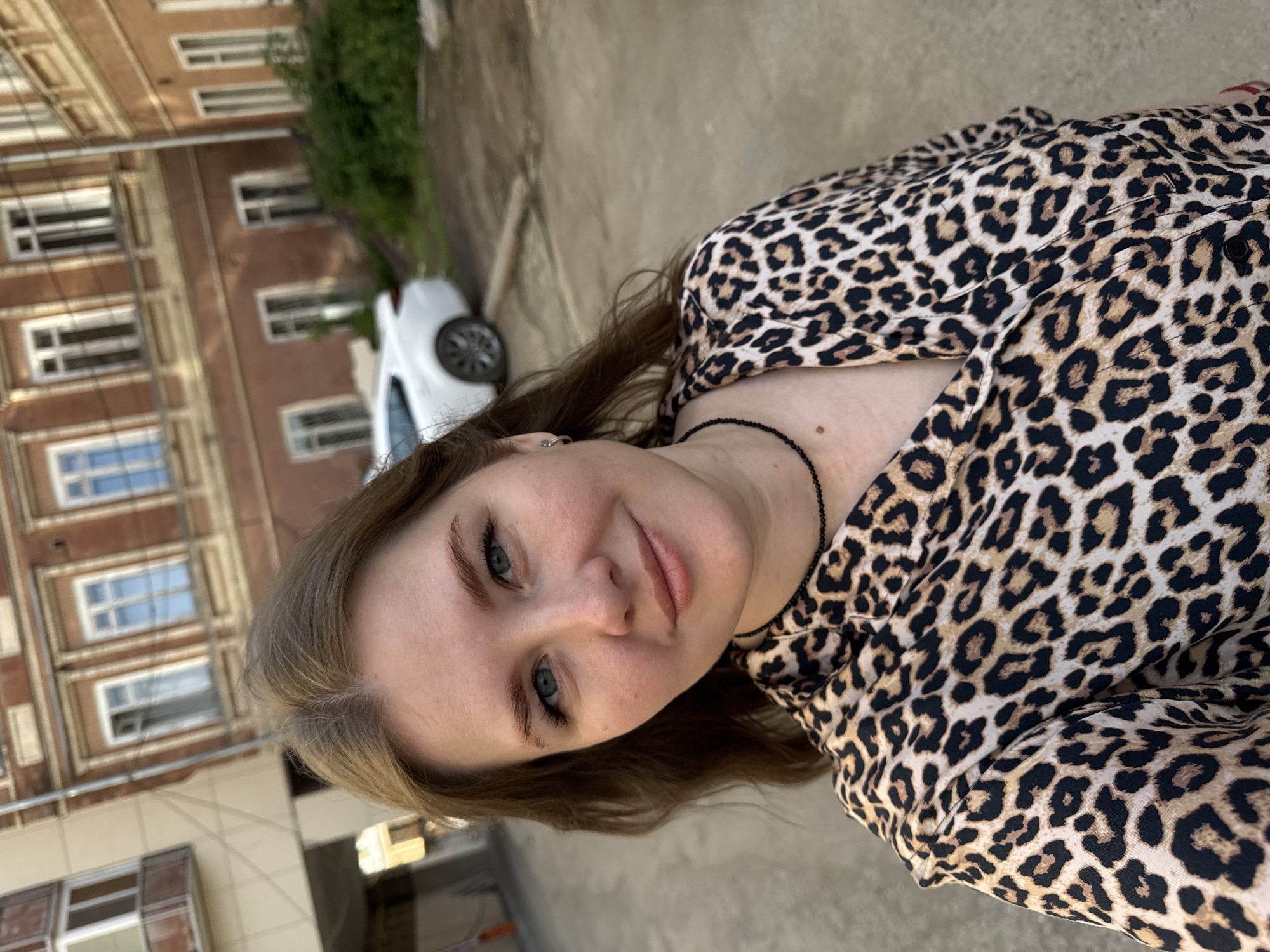 A medium shot shows a woman with light skin, blue eyes, and light brown hair wearing a leopard-print blouse and a black beaded necklace. She is smiling slightly and looking directly at the camera. The location appears to be Perm, Russia, judging by the architecture of the building in the background. The building is made of brown brick with multiple stories of windows. A white car is parked in front of the building. The scene is outdoors, on a paved surface.