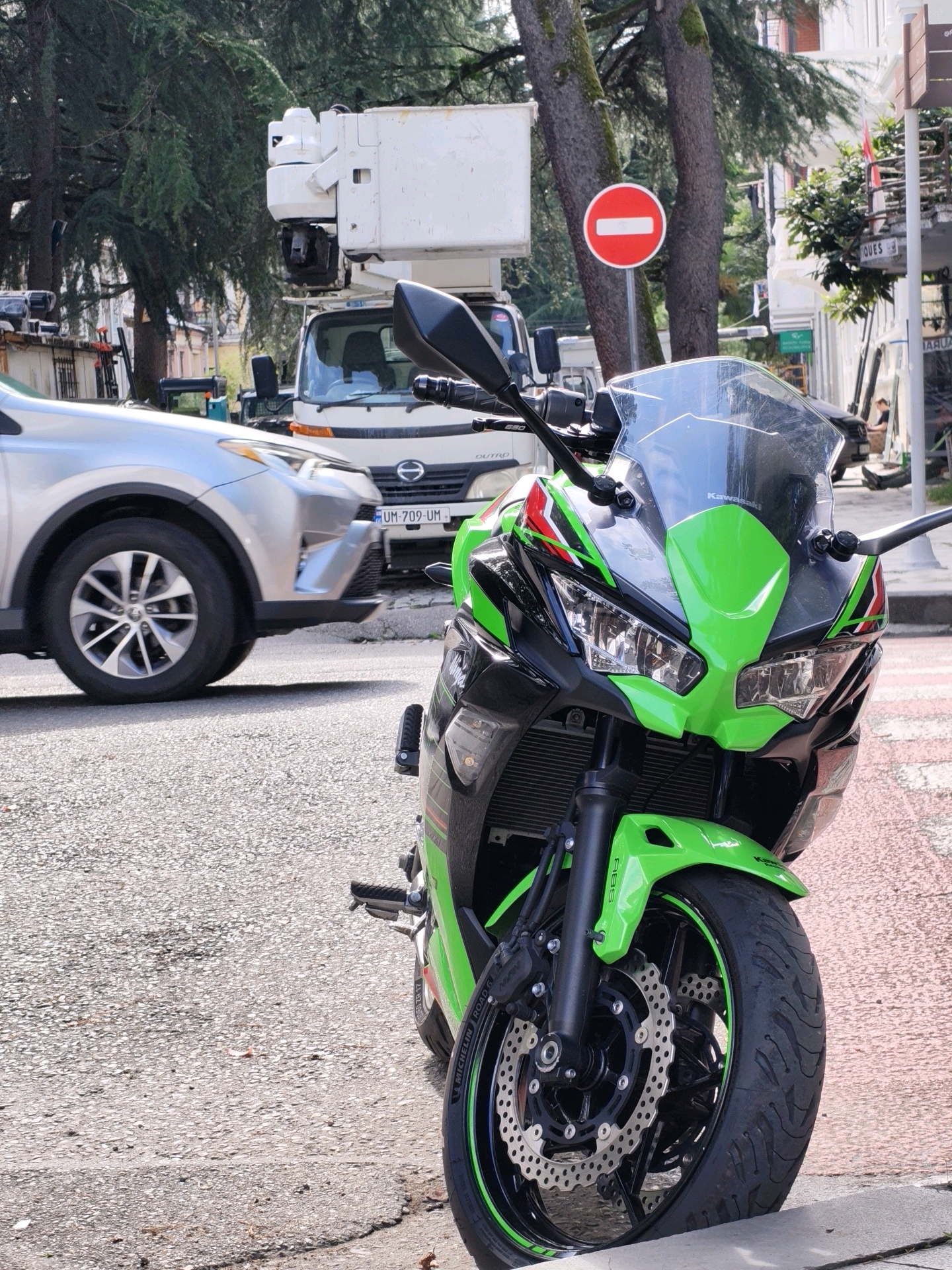 The image captures a street scene in Batumi, Georgia, featuring a bright green and black Kawasaki motorcycle parked on the right side of the frame. The motorcycle is positioned on a reddish paved area, likely a sidewalk or pedestrian zone. In the background, a silver SUV is visible, indicating traffic on the asphalt road. Further back, a white truck with a cherry picker attachment is parked, suggesting possible utility work or construction. A red and white circular "no entry" traffic sign is mounted on a pole behind the motorcycle. Trees with mossy trunks line the street, and buildings with balconies can be seen in the distance, characteristic of Batumi's architecture. The ground is a mix of asphalt and the textured reddish paving.