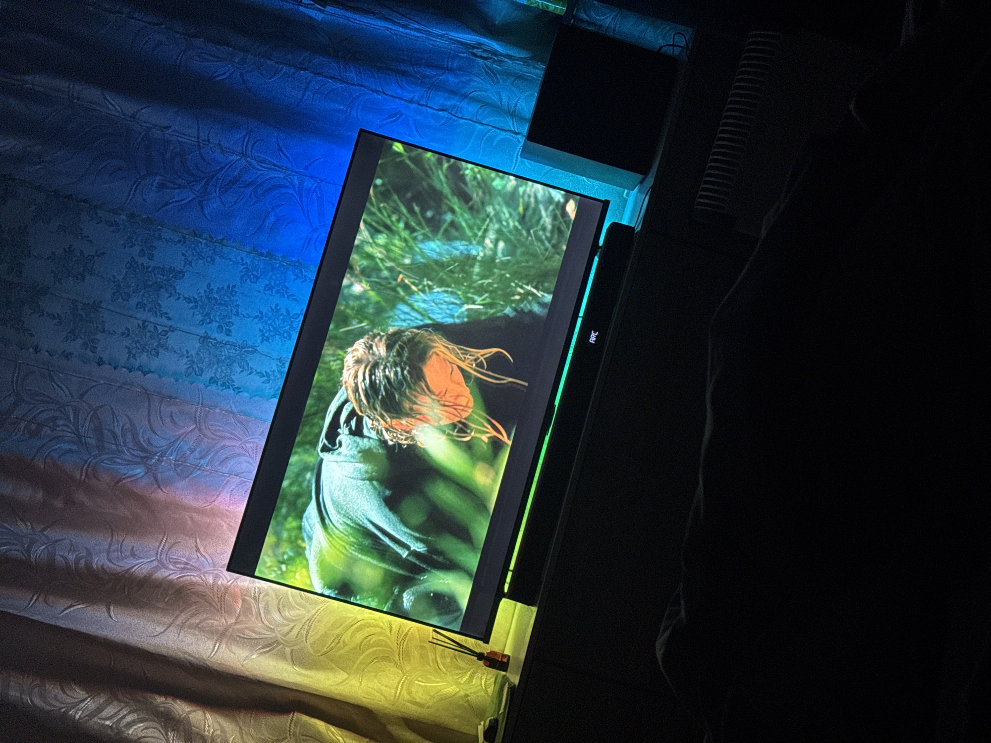 A monitor displaying an image of a person with wet hair and a dark green shirt is resting on a light-colored surface. The surrounding area is dimly lit, with a bluish hue on one side and a dark, shadowy region on the other. A small black object is visible above the monitor. There is no indication of the location being Makiivka, Ukraine.