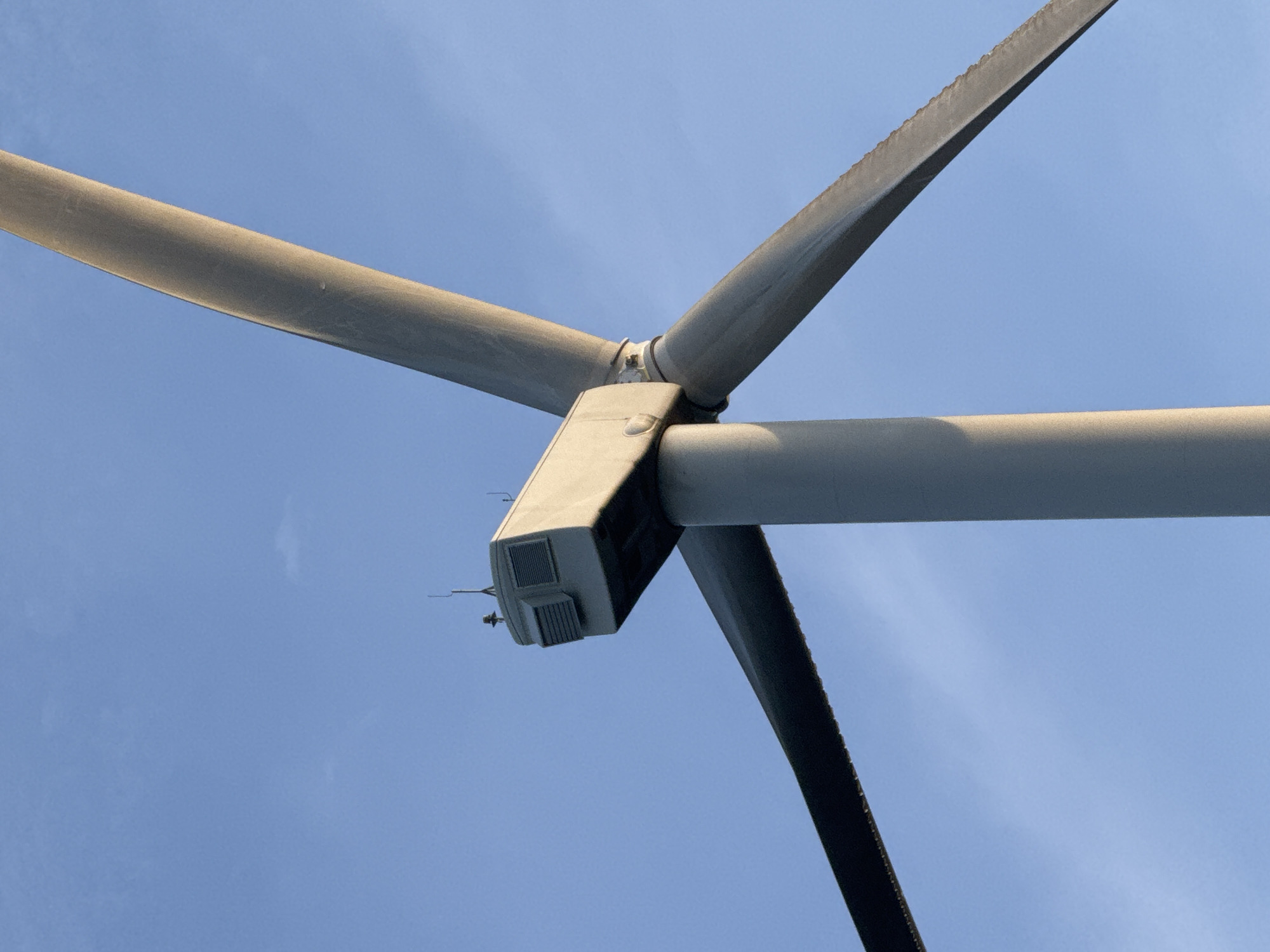 A close-up, low-angle shot captures the nacelle and blades of a wind turbine against a clear blue sky, with a hint of sunlight illuminating one side of the turbine. The nacelle, a grey rectangular structure, is visible at the top, featuring vents on its side and antenna-like protrusions on its roof. Three massive, grey turbine blades extend outwards from the nacelle. One blade is positioned horizontally from left to right, another diagonally from bottom left upwards, and the third extends upwards and to the right, partially cut off by the frame. The scene appears to be located in an open area, likely a wind farm, given the presence of the turbine. While the prompt mentions Phát Chi, Vietnam, the image itself does not contain any overt visual cues to confirm or deny this specific location.