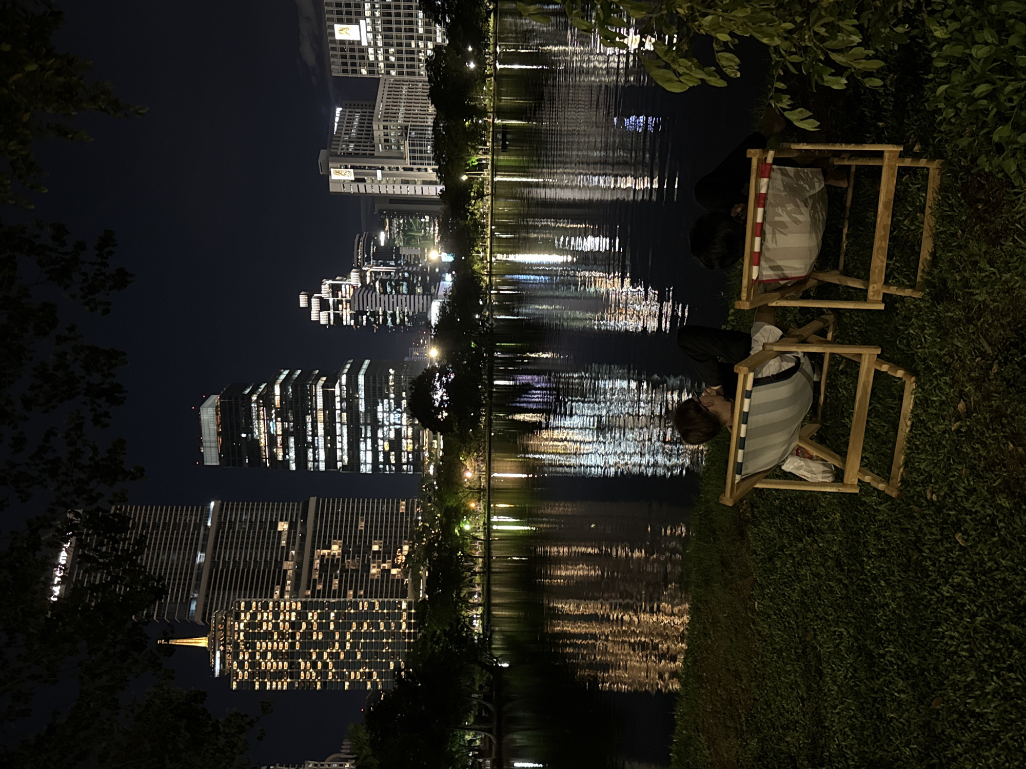 At night, two people sit in Adirondack-style chairs on a grassy bank alongside a lake in Bangkok, Thailand. The illuminated skyline of modern buildings reflects off the water. The chairs are made of light-colored wood and the people are seated facing the water. One person wears a striped shirt, and the other is in dark clothing. The grassy bank slopes down to the water's edge, and green foliage frames the scene. The night sky is dark, and the city lights are bright.