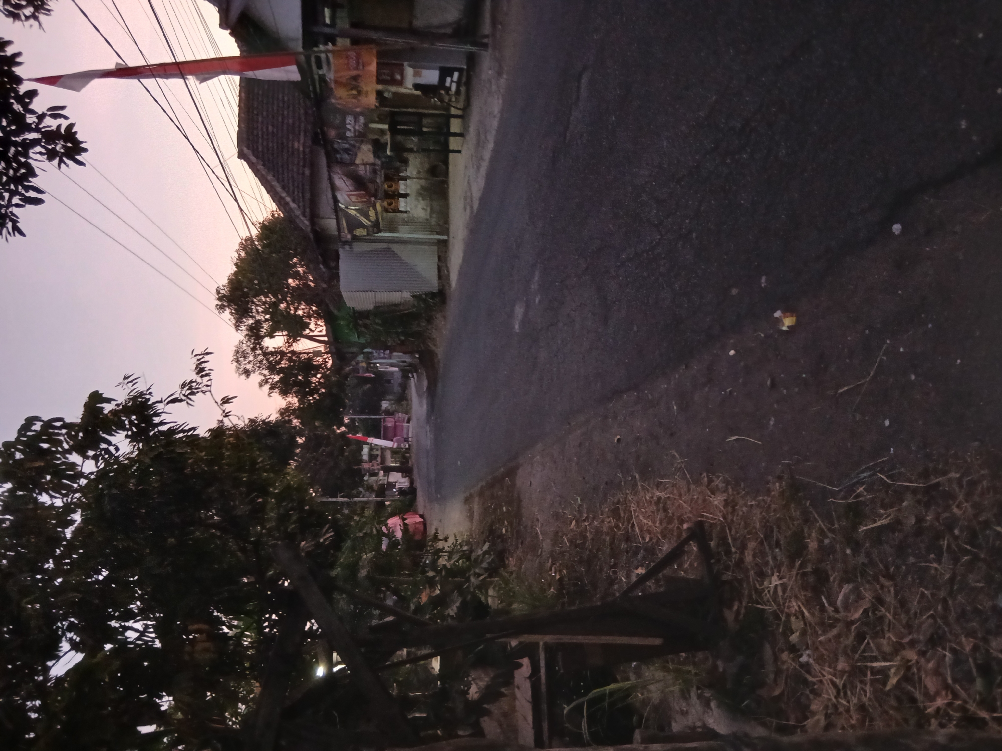 The image depicts a street scene at dusk in Kademangan Kulon, Indonesia. The sky is a soft pink and purple gradient, indicating the transition from day to night. The road, paved with asphalt, stretches into the distance. On the right side of the road, there are several buildings with tiled roofs, and a flagpole with the Indonesian flag (red and white) is prominently displayed. Various advertisements or signs are visible on the building facades. Trees with dark foliage line the left side of the road, casting shadows. Some dry grass and debris are scattered along the side of the road. In the distance, there appears to be a pink vehicle parked on the left side of the street. Power lines are strung overhead, crisscrossing the sky. The overall impression is a quiet, possibly rural or suburban, street at the end of the day.