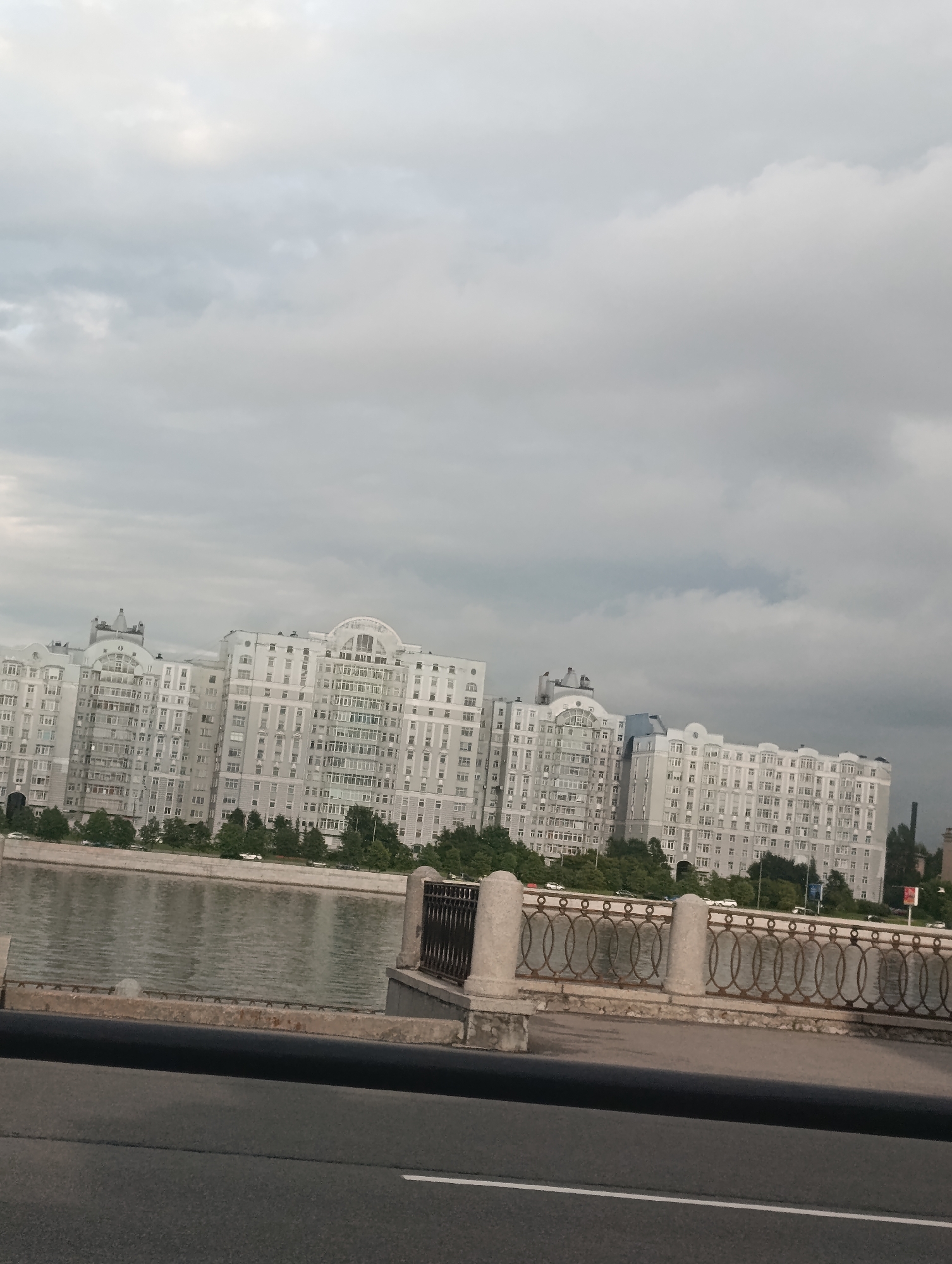 An exterior, eye-level, slightly tilted shot shows a wide, calm river in the foreground, with a large, white, multi-story apartment building situated on the opposite bank. The building features numerous windows and arched elements. A decorative metal railing with oval motifs and stone pillars lines the riverbank in the middle ground. Beyond the building, a dense line of green trees is visible. The sky above is overcast with heavy, gray clouds. The scene appears to be a city landscape in Saint Petersburg, Russia, specifically along the Neva River or one of its branches, given the characteristic architecture and waterfront design. No people are visible in the frame.