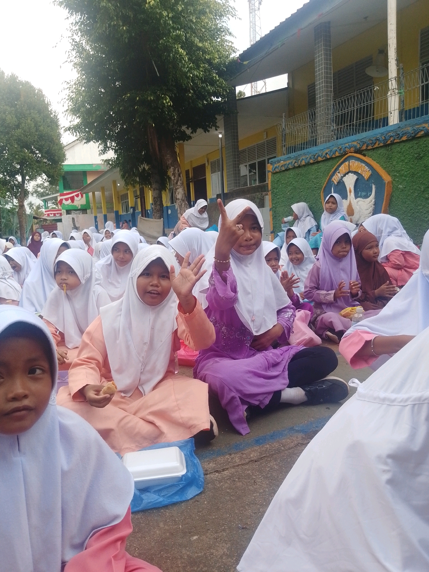 In Batam, Indonesia, a group of young girls, likely students, are gathered outdoors on a paved surface. The girls, dressed in hijabs and modest attire in varying colors, are seated on the ground. Some are holding food, with one girl in the foreground eating. Another girl in a purple outfit is raising her hand, seemingly waving or gesturing. In the background, a building with yellow and green accents and a blue fence suggests an educational institution. Trees with dense foliage are visible to the left, and a tall tower-like structure is present in the distance. The scene appears to be a casual gathering or event, possibly during a break or mealtime.