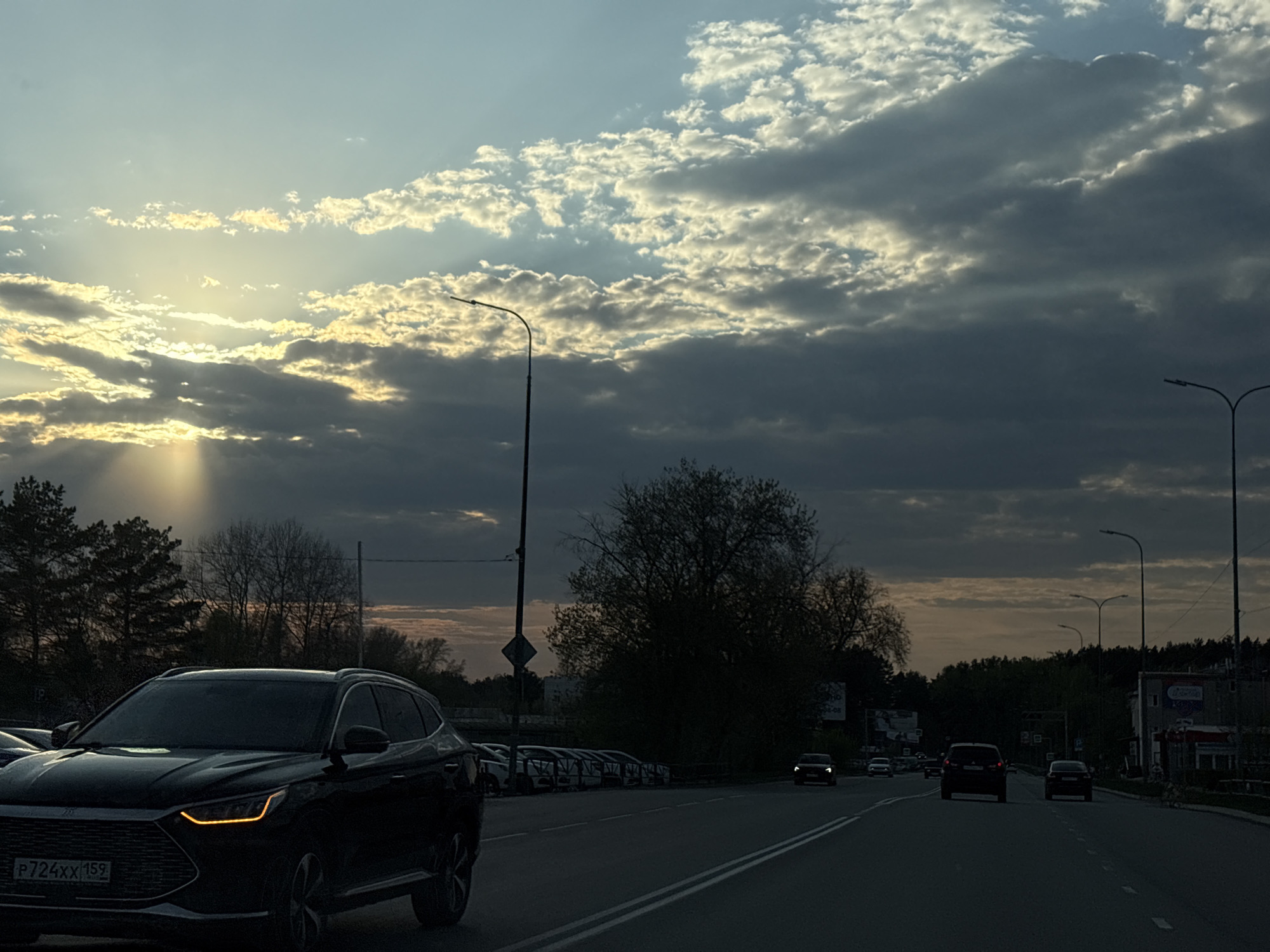The image depicts a street scene at dusk in an unidentified city. The sky is overcast with the sun partially obscured by clouds. A black SUV is in the foreground, traveling along a multi-lane road. Other cars are visible on the road and parked along the side. Streetlights line the road. Trees and buildings can be seen in the background. The license plate on the SUV reads "P724XX 159".