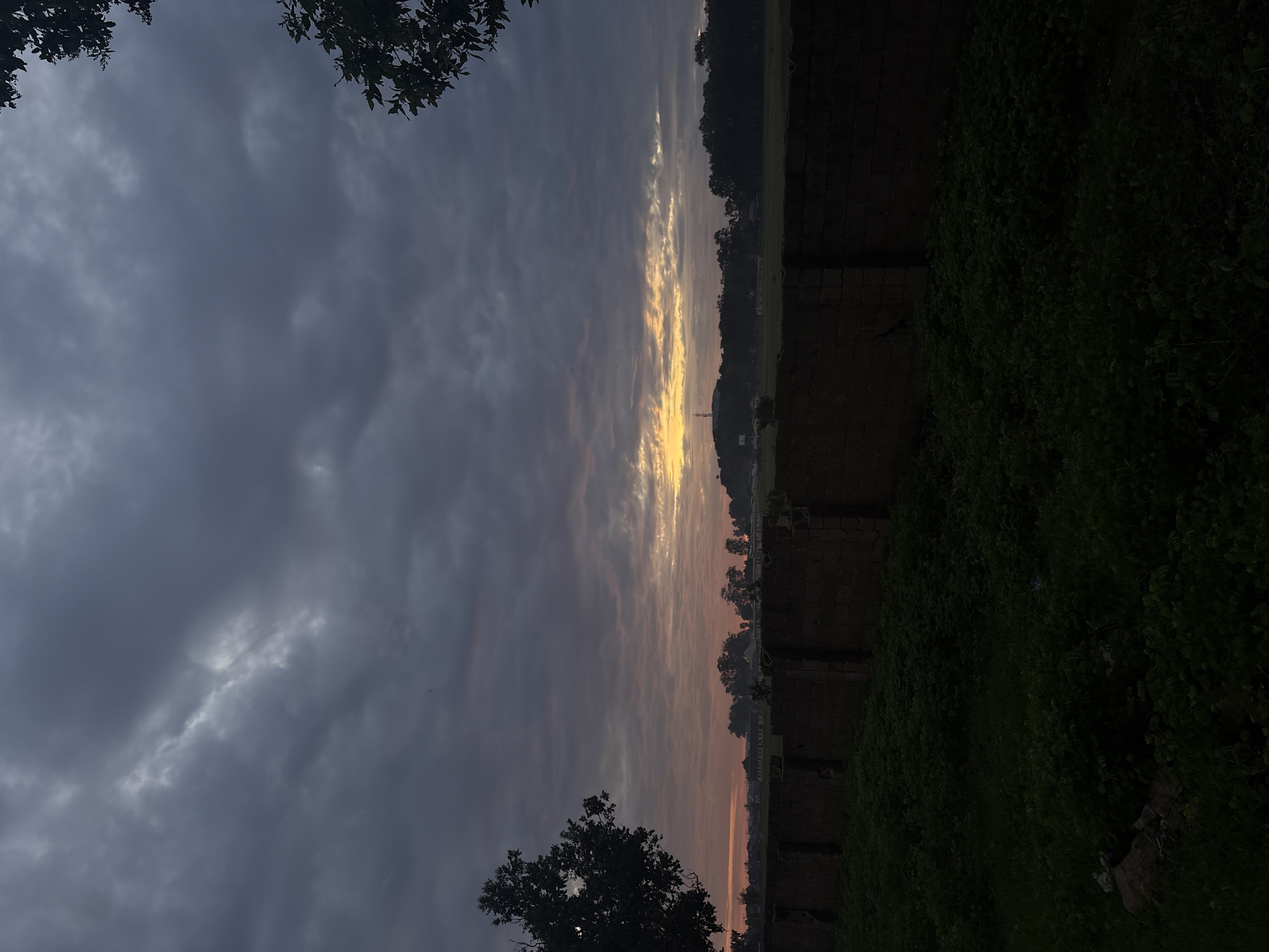 The image depicts an outdoor landscape scene at either sunrise or sunset, situated in the city of Bukuru, Nigeria. The sky occupies the majority of the frame, dominated by a dense formation of dark grey stratus clouds across the upper two-thirds. Below these heavy clouds, a horizontal band of altocumulus clouds is illuminated by a low sun, displaying warm hues of pale orange and pink on the left, transitioning to vibrant yellow and gold towards the center-right.
The horizon line is marked by a continuous, dark silhouette of a tree line extending across the midground. Behind these trees, faint outlines of low-rise buildings are discernible, particularly towards the left. In the center, a small hill or elevated landform rises slightly above the tree line, topped by a slender, dark structure resembling a telecommunications tower or antenna. A large, dark green tree with distinct foliage partially frames the left side of the image, while the leaves of another dark tree are visible entering the frame from the upper right corner.
In the foreground, a dark, low-lying masonry wall, constructed from rough, dark brown bricks or blocks, spans horizontally across the lower third of the image. The wall appears somewhat weathered or unfinished. Immediately in front of and below the wall, the ground is covered with a thick, dark green carpet of dense foliage, consistent with overgrown grass and low-lying shrubs. No human figures, animals, or explicit human activity are visible within the frame.