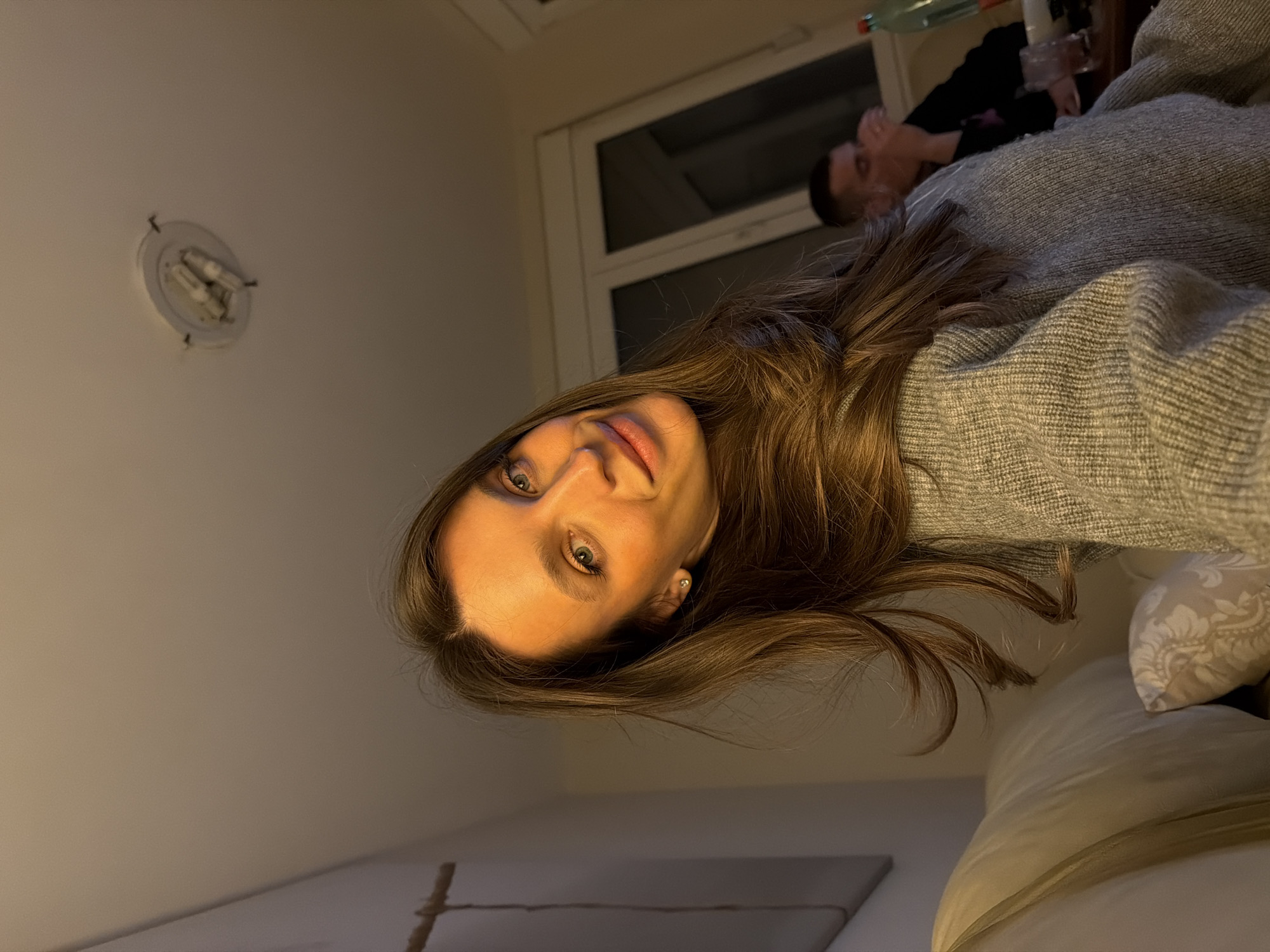 A person with long brown hair wearing a gray sweater poses in a bedroom with a white wall and ceiling light. In the background, another person is visible near a window. The location is likely indoors, but the city of Budva, Montenegro, cannot be verified from the provided image. Pillows and a bed are also visible.