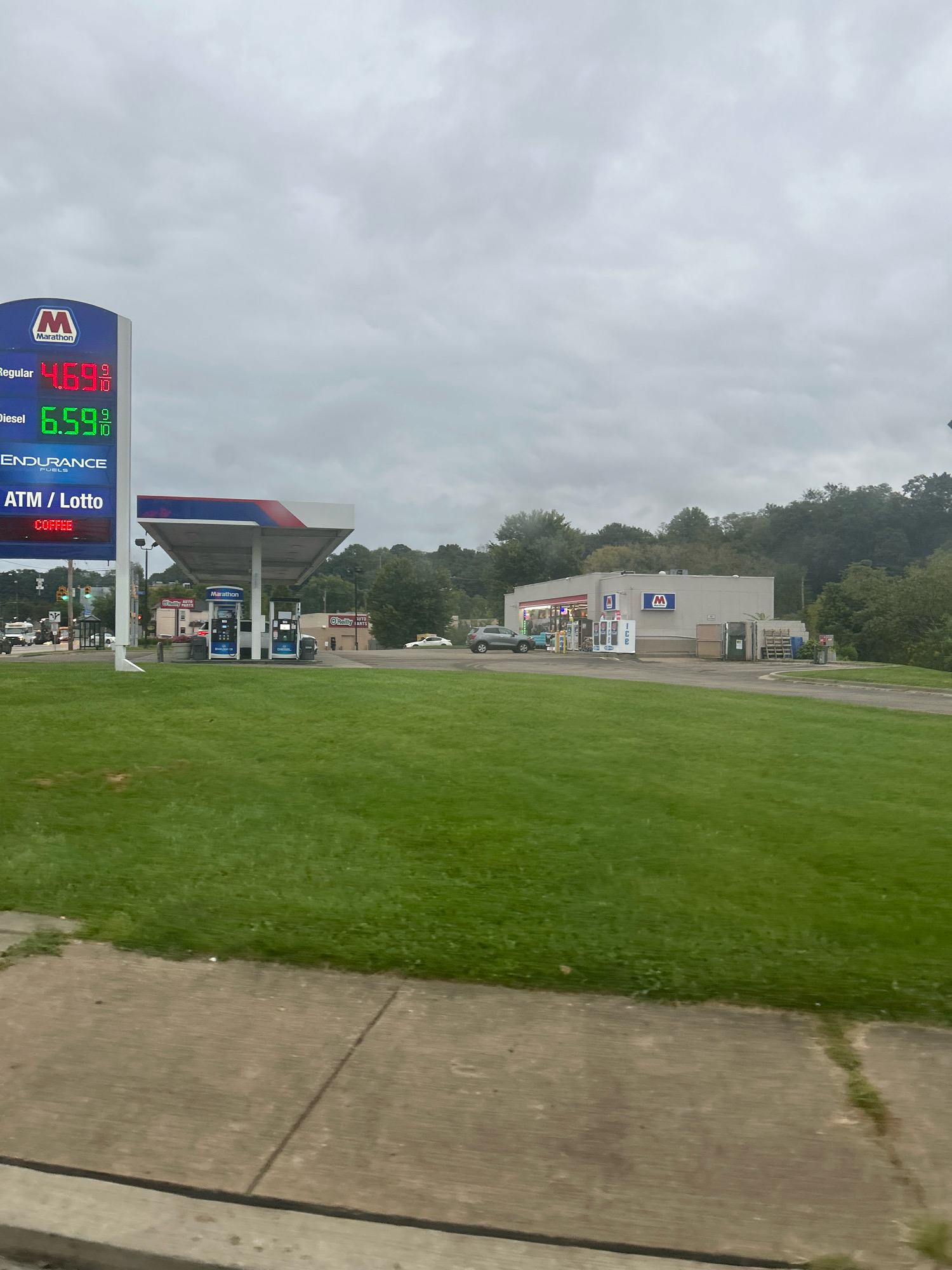 An exterior, eye-level, medium shot captures a Marathon gas station on an overcast day. The gas price sign on the left displays "Regular 4.69 9/10" and "Diesel 6.59 9/10". Below that, it reads "ENDURANCE FUELS" and "ATM / Lotto COFFEE". The gas station features a covered area with gas pumps. To the right of the gas station is a convenience store with a Marathon logo. A white car is parked at a gas pump, and a grey car is visible to the left of the store. Across the street, another gas station with a "Rally" sign is visible. The area is surrounded by green grass and trees in the background. The image appears to be taken from a vehicle driving by.