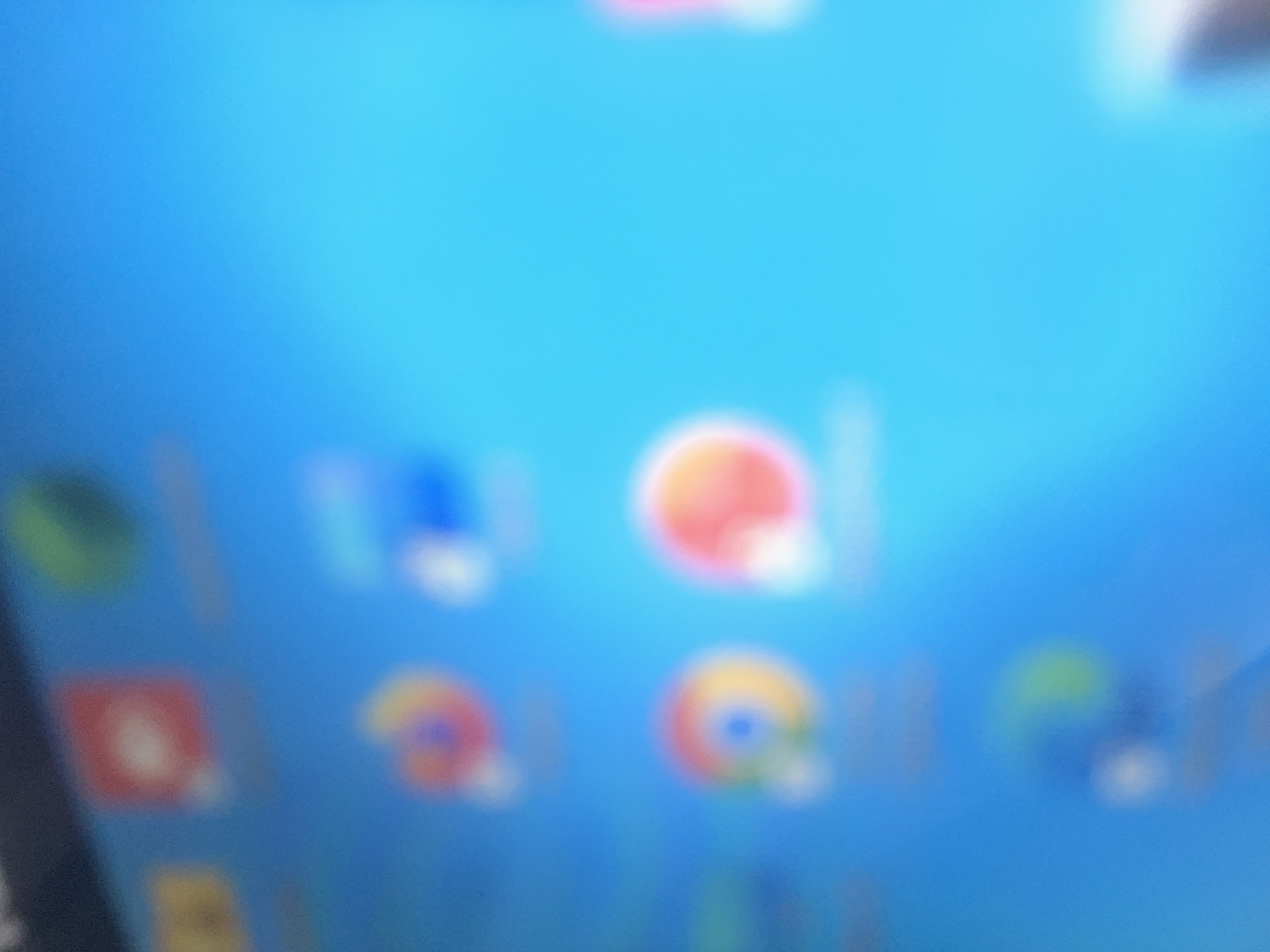 The image shows a blurred view of a computer or tablet screen. The background is a light blue color. There are several blurry icons visible, including what appear to be the icons for Google Chrome, other Google apps, and potentially other app icons. The icons are arranged in a grid pattern. The image is out of focus, making it difficult to identify details precisely. The image does not contain any information pertaining to the city of Dar es-Salaam, Tanzania.