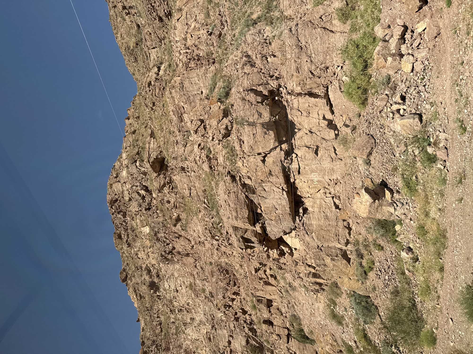 A low-angle shot captures a steep, rocky mountainside under a clear blue sky. The mountain is composed of large, rough-hewn rocks in shades of brown and tan, with some sparse green vegetation clinging to its surface. A thin white wire is visible stretching across the sky diagonally from the upper right to the upper left. The ground in the foreground is covered with dirt, gravel, and more vegetation, suggesting a natural, rugged landscape. The scene appears to be a remote, mountainous area, possibly on the outskirts of Krasnyy Most, Kyrgyzstan, given the context. There are no people or animals visible.