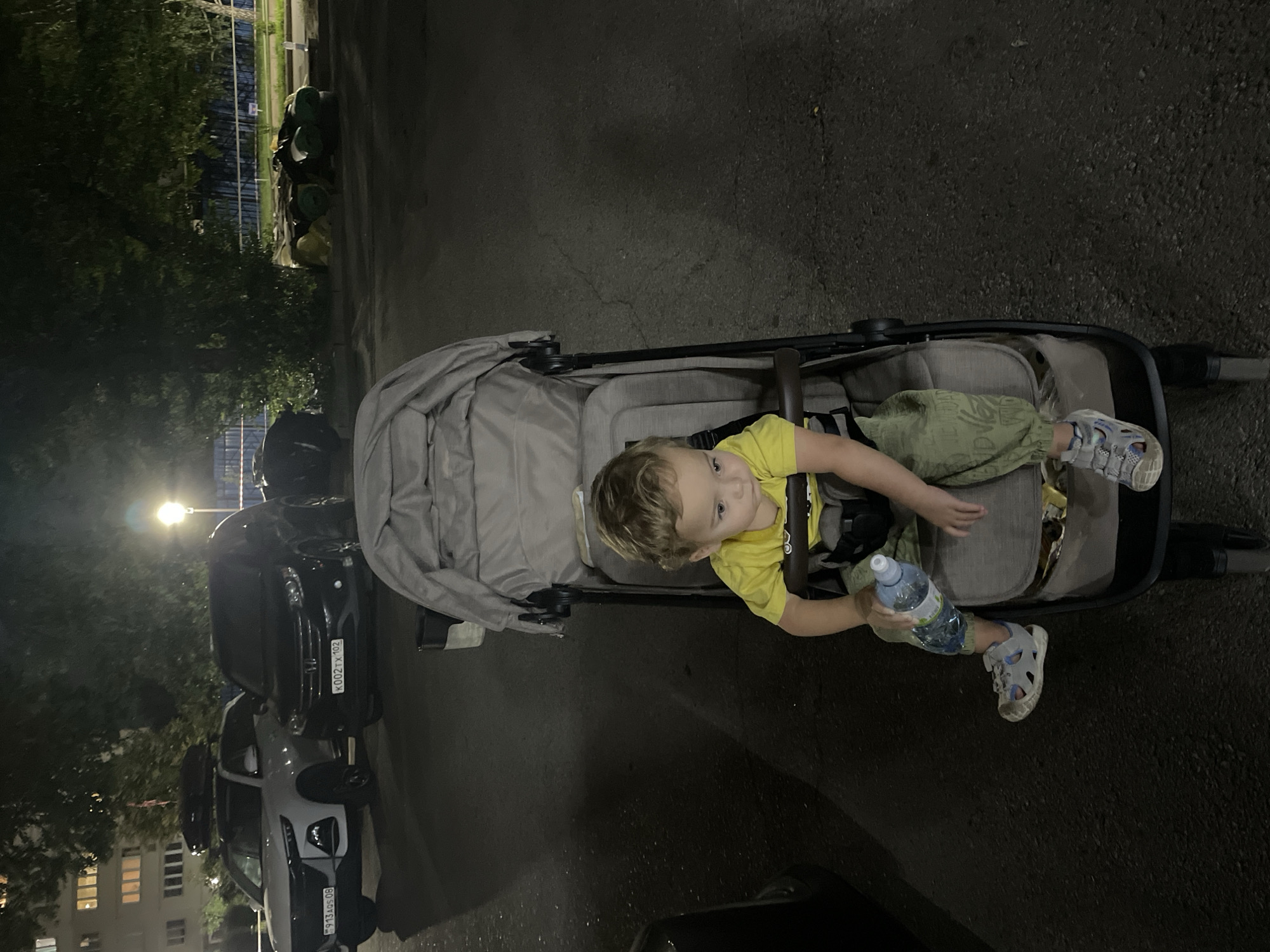 The image shows a toddler sitting in a stroller outdoors at night. The toddler is wearing a yellow t-shirt and green pants, and is holding a bottle of water. In the background, there are several cars parked on asphalt. A building with lit windows is visible on the left, and trees with dark foliage are on the right. The scene appears to be in a residential area or parking lot, possibly in Almaty, Kazakhstan, given the presence of Cyrillic license plates on the vehicles.
