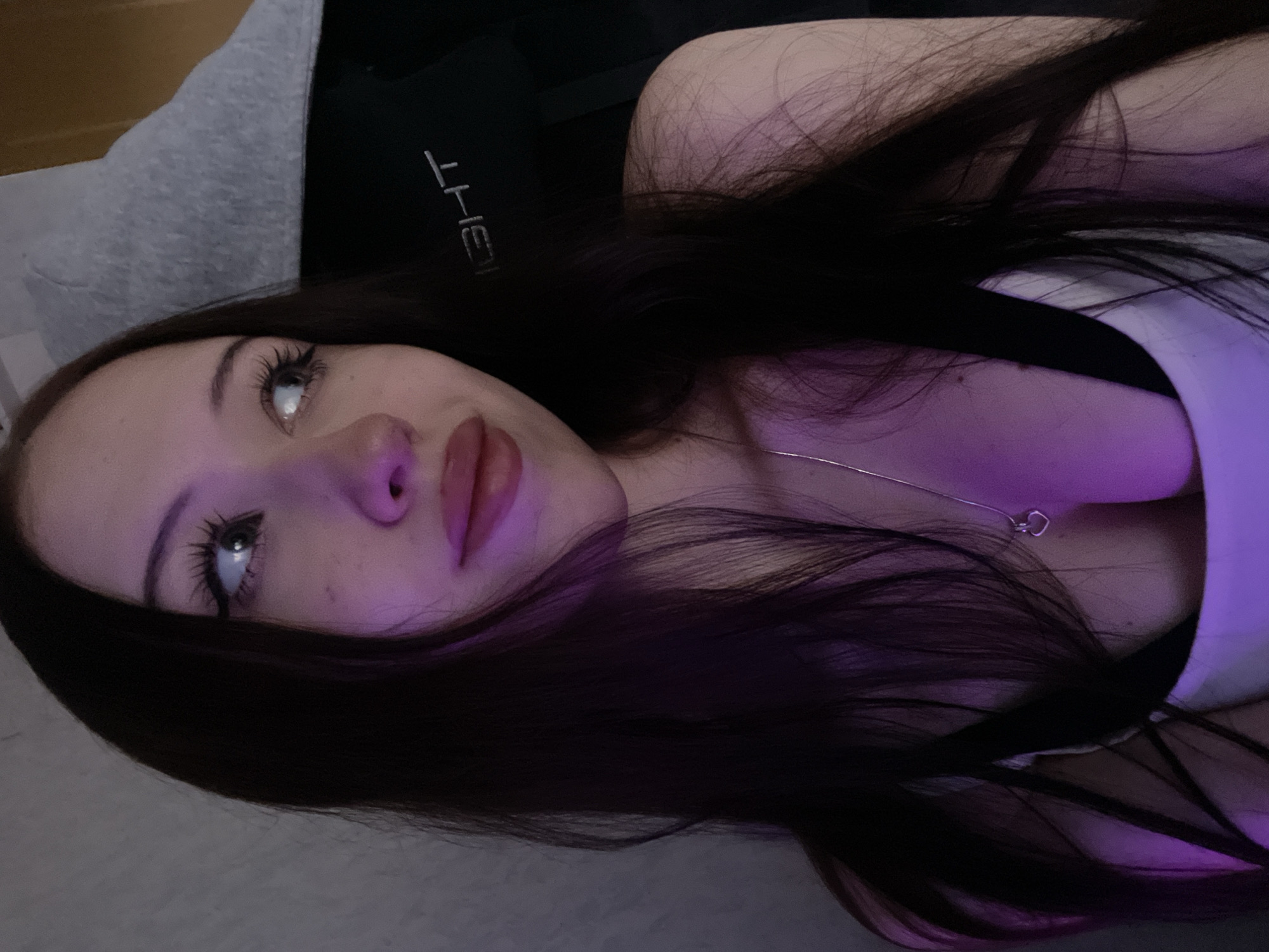 The image shows a close-up of a young woman with long, dark hair. She is looking to the right of the frame, and her lips are slightly parted. She is wearing a white top with black straps and a silver necklace with a heart-shaped pendant. A purple light is cast on the left side of her face and hair. In the background, a dark chair with white lettering that appears to read "LIGHT" is visible. The scene suggests an indoor setting, likely a room in Votkinsk, Russia, possibly a home or a studio, with ambient lighting that includes a colored light source.