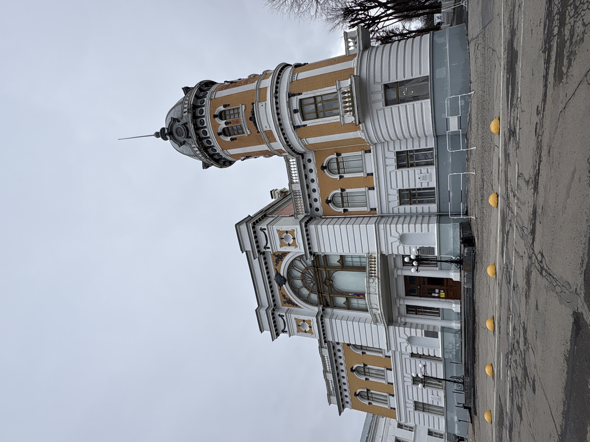 The image displays a grand, ornate building under an overcast sky. The multi-story structure exhibits a distinctive architectural style, characterized by a blend of Neoclassical and possibly Art Nouveau elements. Its facade features alternating sections of light brown brickwork and white architectural details, including fluted pilasters, cornices, balustrades, and decorative elements around windows and entrances.
A prominent circular tower rises on the left side of the building, echoing the primary color scheme. This tower is adorned with multiple windows and topped by a domed roof culminating in a slender spire. The main body of the building has a variety of window shapes, including arched and rectangular frames, some with decorative surrounds. The central entrance features a large, ornate arched window or glass canopy above the main doors, which are flanked by decorative lampposts.
In front of the building is an asphalt ground surface, possibly a street or parking lot, marked with white lines and dotted with several yellow hemispherical bollards. The asphalt appears worn with visible cracks. In the upper right background, bare tree branches are visible against the cloudy sky, suggesting a leafless season, likely winter or early spring.
The sky is a uniform light grey, indicating an overcast day with muted daylight. No people or specific activities are depicted; the scene is focused solely on the architecture. Inside one of the large arched windows, a small flag with vertical red, white, and blue stripes is visible, likely indicating a Russian flag, which aligns with the Ulyanovsk, Russia context. Small, illegible notices are also visible on the main entrance doors.