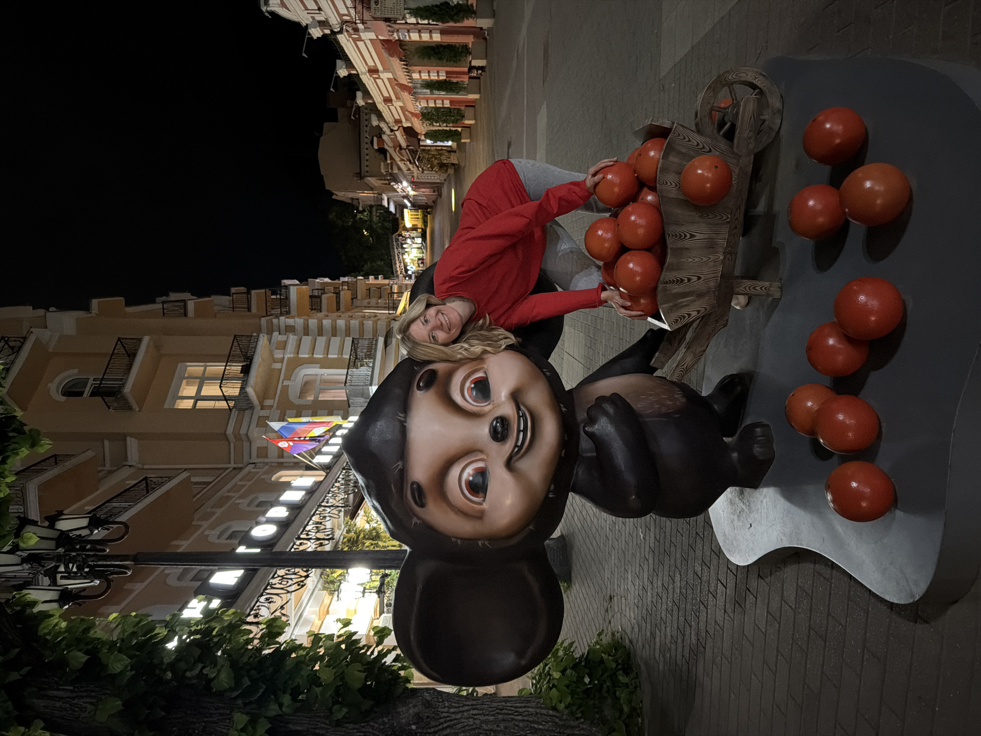 A blonde woman in a red sweatshirt and grey pants poses with a statue of Cheburashka, a fictional character, in the city of Kislovodsk, Russia. The statue is large and dark brown with big ears and red eyes. The woman is leaning down, placing her hands on a small wooden wheelbarrow filled with bright red, round objects that appear to be decorative tomatoes. Several more of these red objects are scattered on the ground in front of the statue and wheelbarrow. The scene is set at night on a paved sidewalk lined with trees and buildings. A streetlamp with multiple lights is visible to the left of the statue. The buildings in the background have balconies and decorative architectural features, illuminated by lights. Flags, including the Russian flag, are visible on one of the buildings. The overall impression is a nighttime street scene in a resort town.