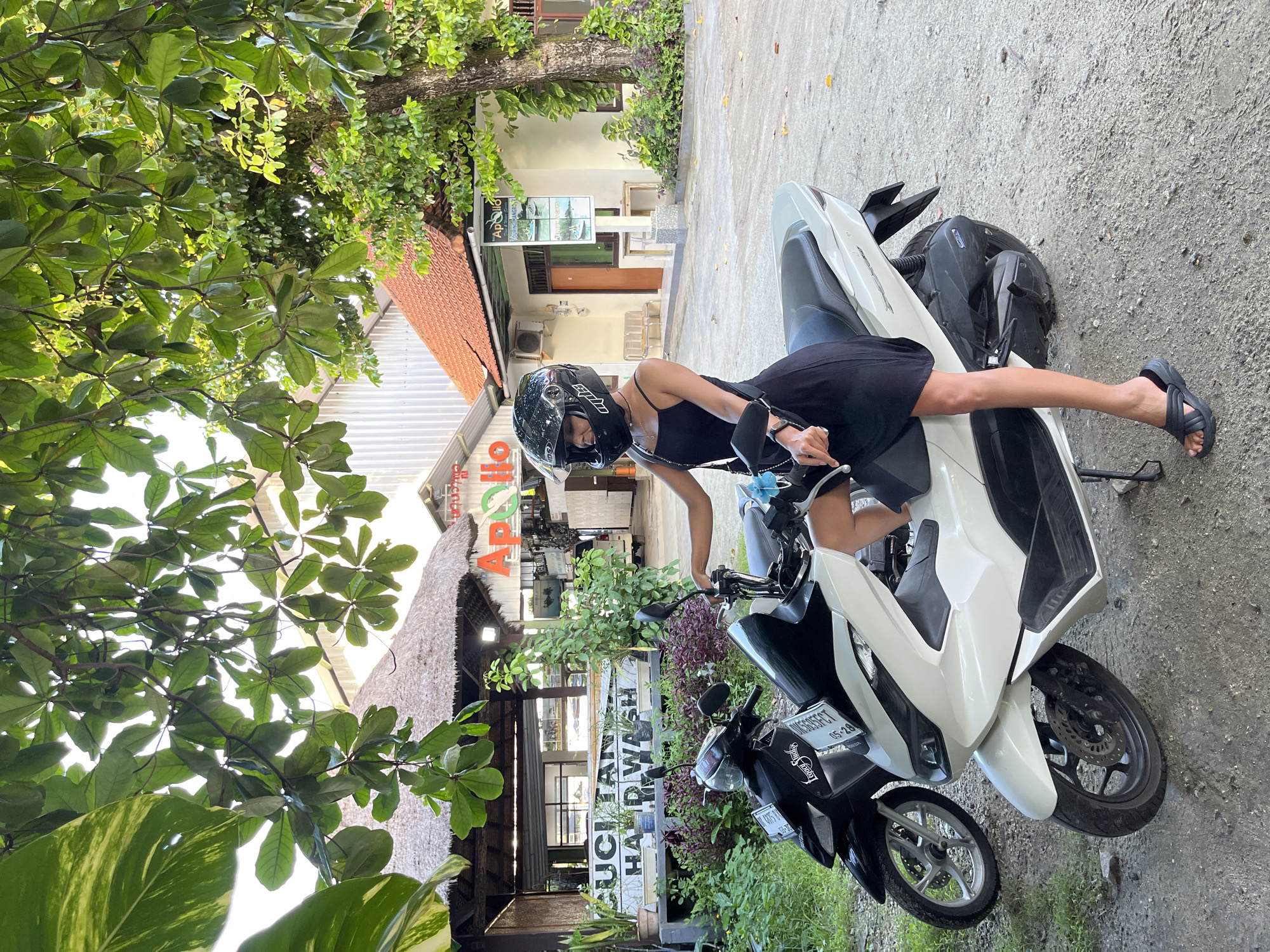 The image is presented in a portrait orientation, rotated 90 degrees clockwise, depicting a woman on a scooter in a bright, outdoor setting. The ground appears on the right side of the frame, and the sky and treetops on the left.
The central subject is a woman, smiling and wearing a black full-face helmet (with "SU" visible on the side) and a black sleeveless dress. She is seated or posing on a white and black scooter, holding the handlebars. Her left leg is extended towards the ground. The scooter's front fender and bodywork are white, while the seat and rear components are black. A license plate on the front of the scooter is clearly visible, reading "DK6505ECT 05-28", which is a Bali registration. A second, dark-colored scooter is partially visible behind the main scooter, with a license plate starting "DK 4851 I...".
The background is rich with tropical foliage, including large green leaves framing the scene from the left side (originally top). Bright sunlight filters through the dense canopy. Behind the scooter and woman, various buildings line what appears to be a street or path. One building has a distinctive terracotta-tiled roof, while another features a rustic thatched roof. A sign on one building clearly displays "APollo", likely a business name. Further back, another sign partially reads "KUCIAH HANDWASH", suggesting a hand-wash service. The ground on the right of the image is paved with concrete or asphalt. The scene takes place during the day, under clear and bright conditions in Kuta Selatan, Indonesia.