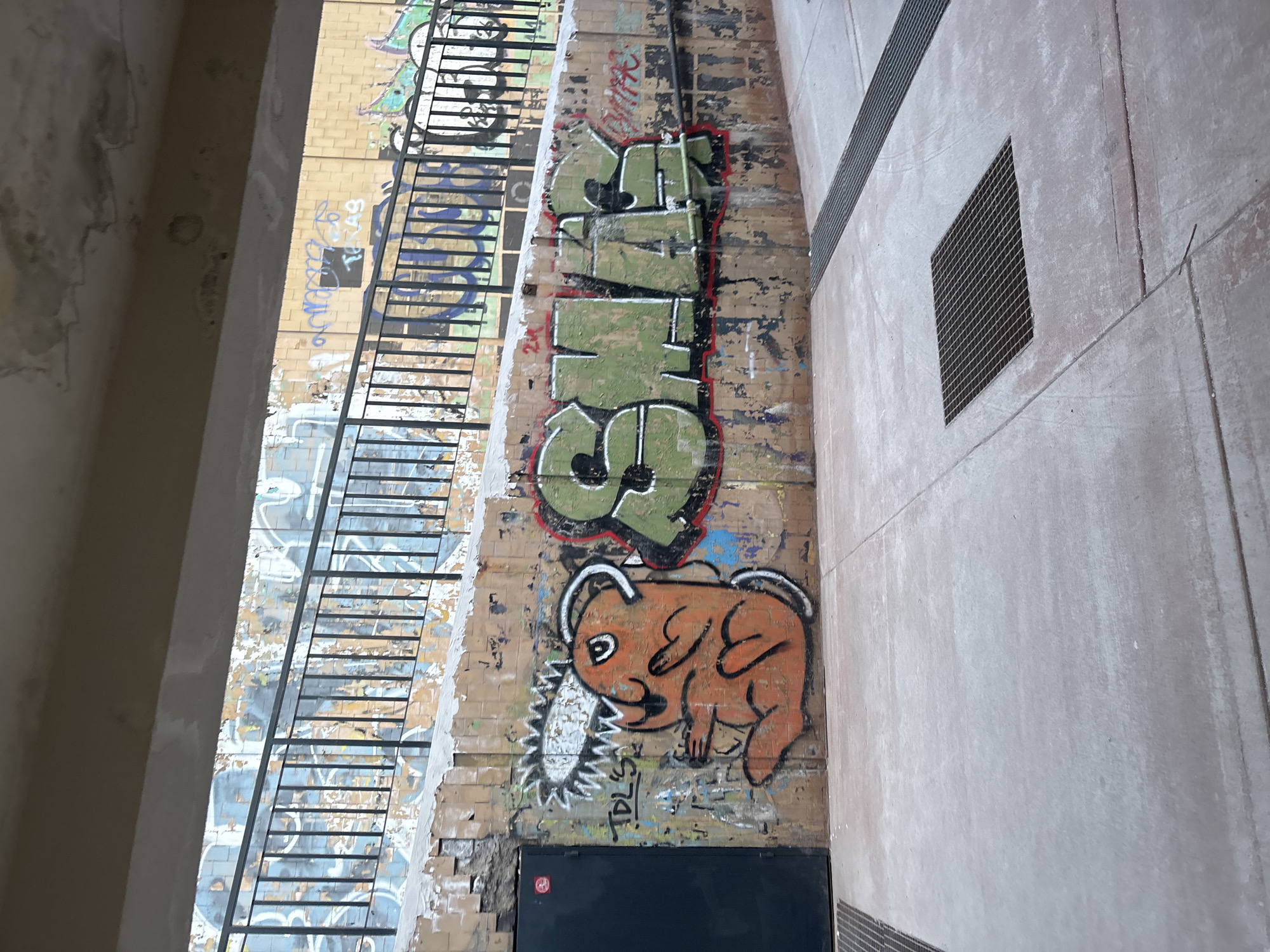 The image displays a graffiti-covered pedestrian underpass or tunnel in Sofia, Bulgaria. A weathered concrete ceiling beam, exhibiting peeling paint and discoloration consistent with water damage, frames the top of the scene. Below, an inclined wall, constructed from light-colored tiles, is largely obscured by numerous layers of spray paint and graffiti. This upper wall is fronted by a black metal railing with vertical bars. The lower portion of the scene reveals a light beige-tiled retaining wall, also heavily marked with graffiti. On the far left of this lower wall, a dark, recessed metal door bears a red circular sticker with a white symbol. Adjacent to the door, a prominent orange cartoon character, resembling Pochita from Chainsaw Man with a chainsaw-like head, is spray-painted. To the right of this character, large green block letters with red outlines spell "SMAG". The concrete floor in the foreground is light gray, showing signs of wear, and contains two metal grate drains: a square one centered in the frame, and a longer, linear one extending along the right edge. No individuals or specific activities are visible within the scene.