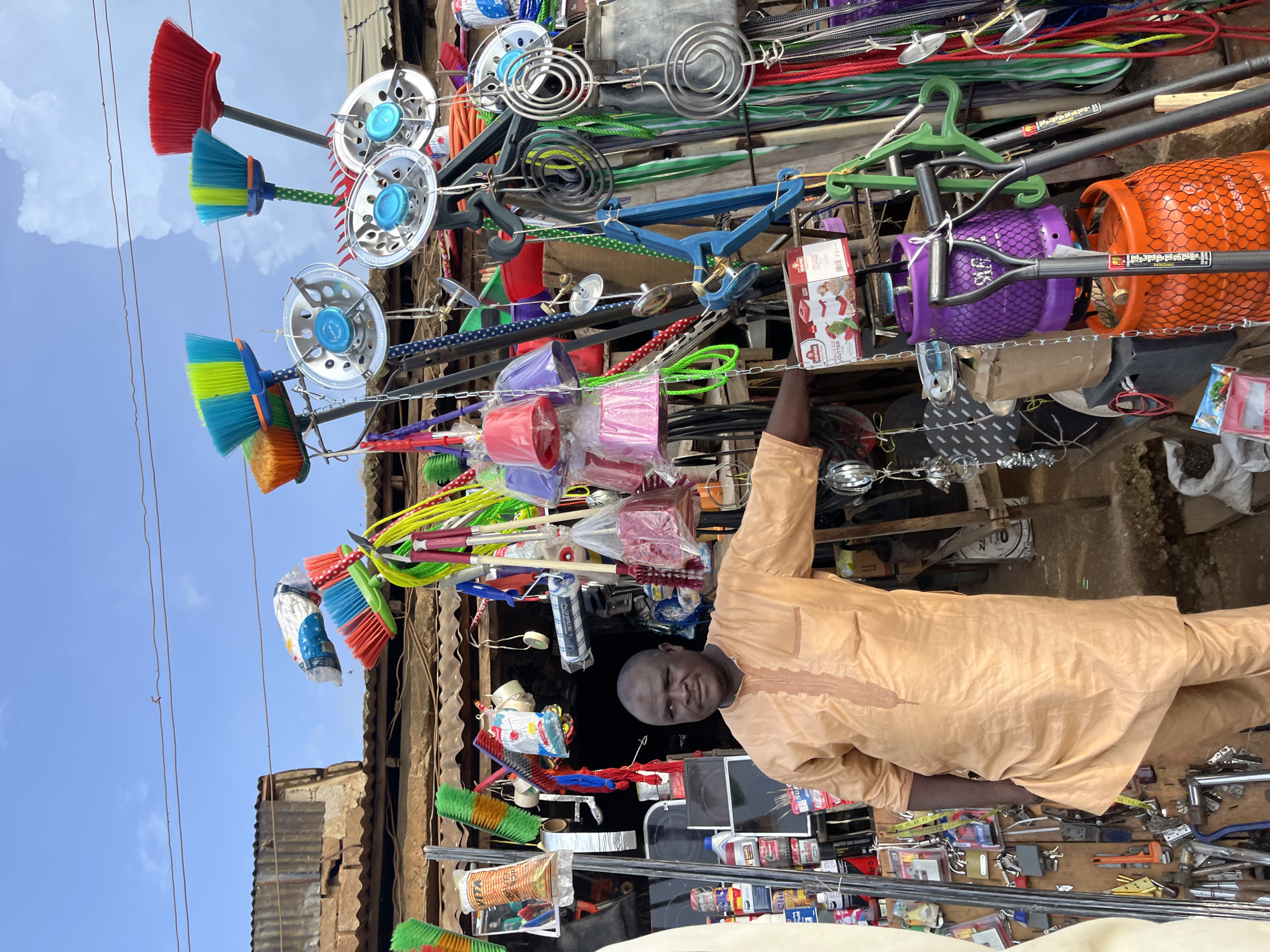 In Rafin Par, Nigeria, a man stands in front of a storefront displaying an assortment of household goods. The man, dressed in a light orange tunic and pants, has a neutral expression and is positioned slightly to the left of the frame, with his left arm resting on his hip. The storefront is cluttered with items such as brooms with colorful bristles, gas cylinders, various kitchen utensils, cleaning supplies, and other miscellaneous objects hanging from the ceiling and displayed on shelves. The background reveals a rustic, open-air market setting with corrugated iron roofing and exposed wooden structures, suggesting a typical local marketplace environment. The sky above is blue with scattered clouds.