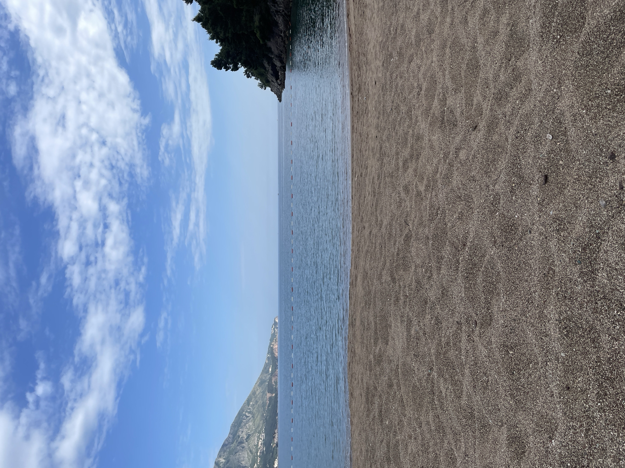 The image depicts a beach in Bečići, Montenegro, with a strip of dark sand on the right and blue water separated by a safety rope. On the left, a blue sky with scattered clouds is visible. A rock formation covered with greenery juts into the sea in the upper center, and another rocky, mountainous area is visible in the lower left.