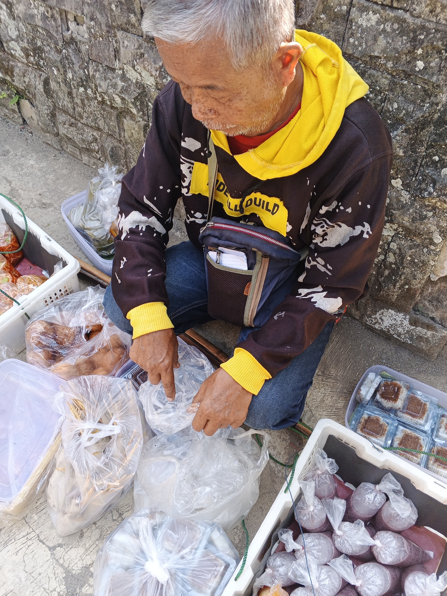 A man is sitting on the ground in front of a stone wall in Warungkiara, Indonesia, surrounded by plastic bags and containers of food items. He is wearing a dark hooded sweatshirt with yellow accents and blue jeans. His hands are visible, and he appears to be sorting or arranging the food. The food items include what look like packaged snacks in a clear container, and in other bags, there are what appear to be fruits or drinks in smaller plastic bags. The ground is paved with concrete, and there is a green rope visible. The overall scene suggests a street vendor preparing or displaying their goods for sale.