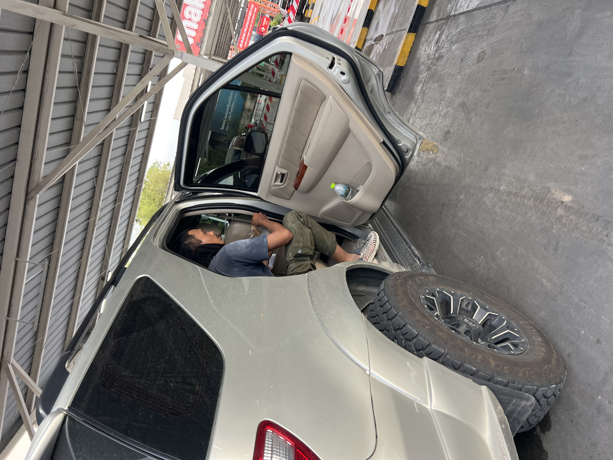 A man is reclining in the driver's seat of a silver Toyota Fortuner. The driver's side door is open. The vehicle is parked under a metallic canopy, possibly at a commercial establishment. The background hints at a commercial area. The tire tread and wheel design are clearly visible. The location is Baan Bo Phut, Thailand.