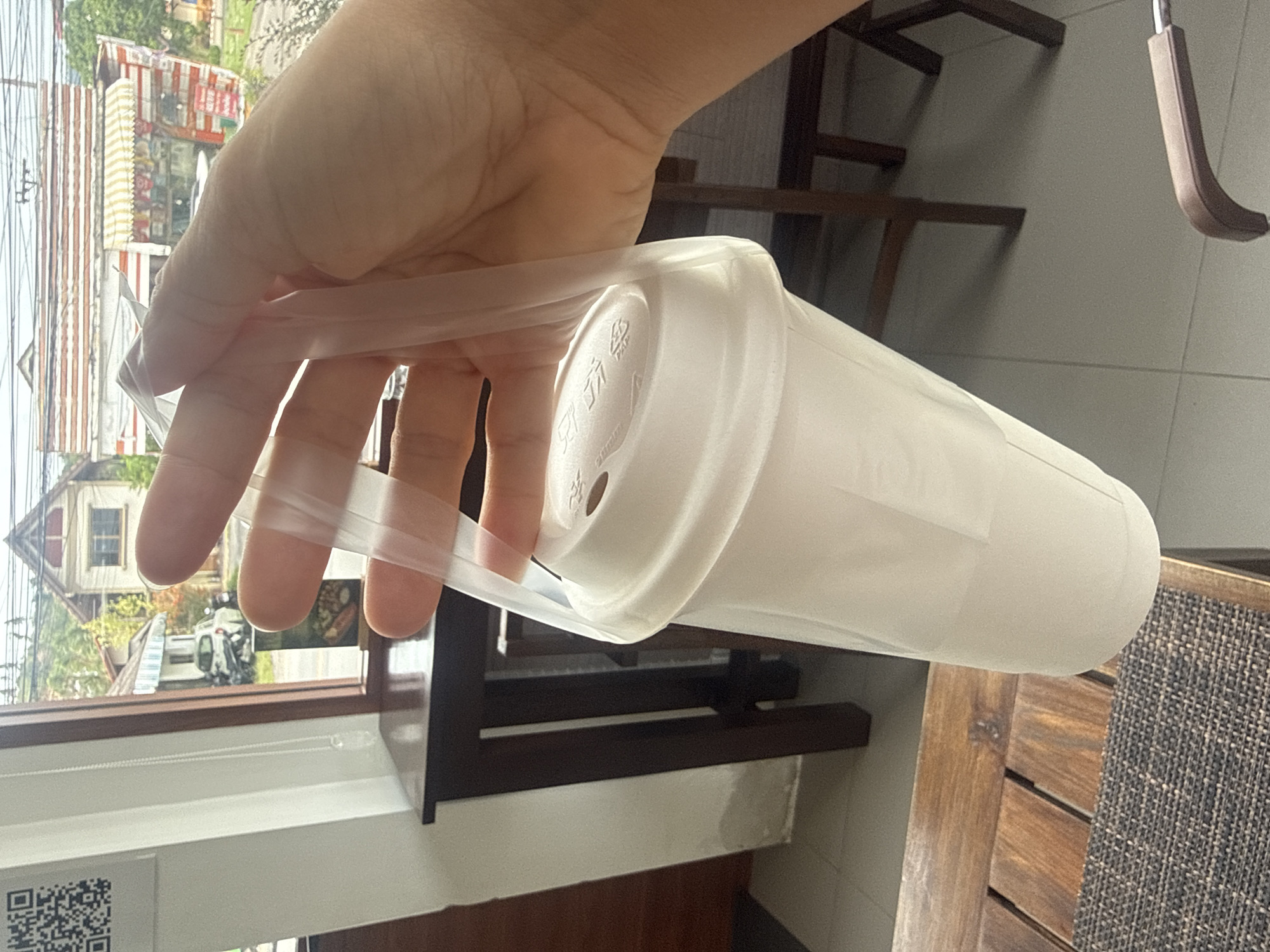 A person's hand holds a clear plastic bag containing a large white disposable cup with a white lid. The cup has a small circular opening on the lid. The bag is folded over the top of the lid and held by the person's thumb and index finger. The background shows a view through a window of a street scene in Ko Samui District, Thailand. A white car and a motorbike are parked on the street in front of a building with a striped awning. The scene appears to be indoors, likely a cafe or restaurant, with a wooden table and chairs visible in the foreground. The lighting suggests it is daytime.