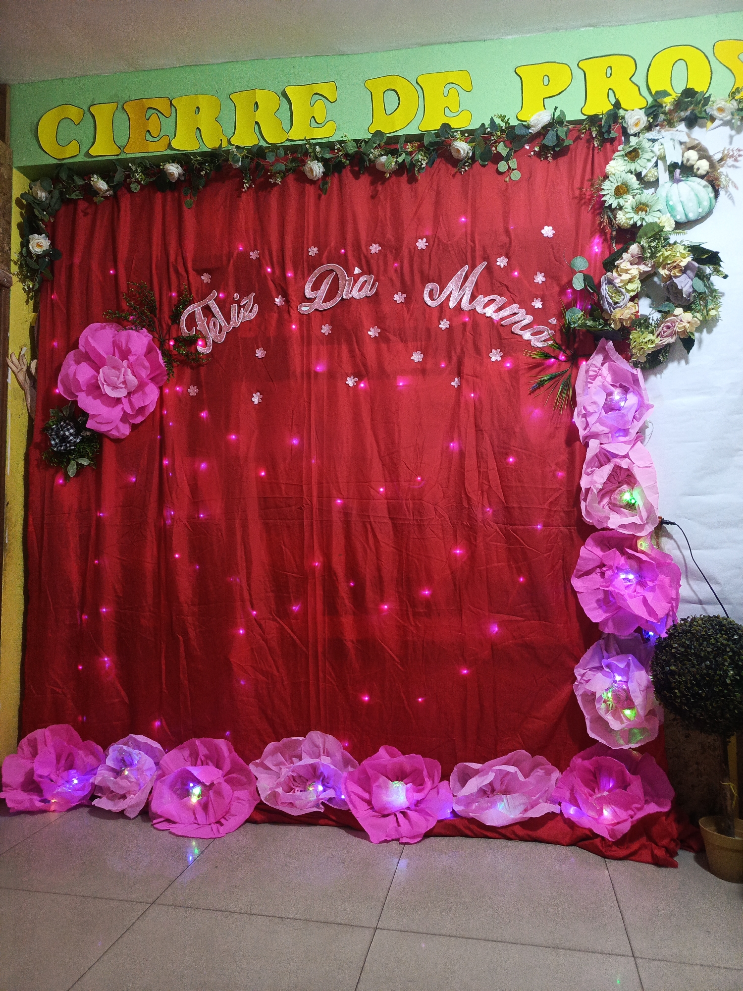 The image shows a decorated backdrop, likely for a celebration or event, set against a wall and floor that suggests an indoor setting. The backdrop consists of a red fabric with small, twinkling lights, the words "Feliz Día Mamá" written across the center, and surrounded by flower decorations of various colors and sizes. The words "CIERRE DE PRO" are visible above the fabric, potentially indicating the event is a "closure" or final celebration for a program. The room has tile flooring and a light-colored wall. The image provides no information about the city, Juan Griego, Venezuela.
