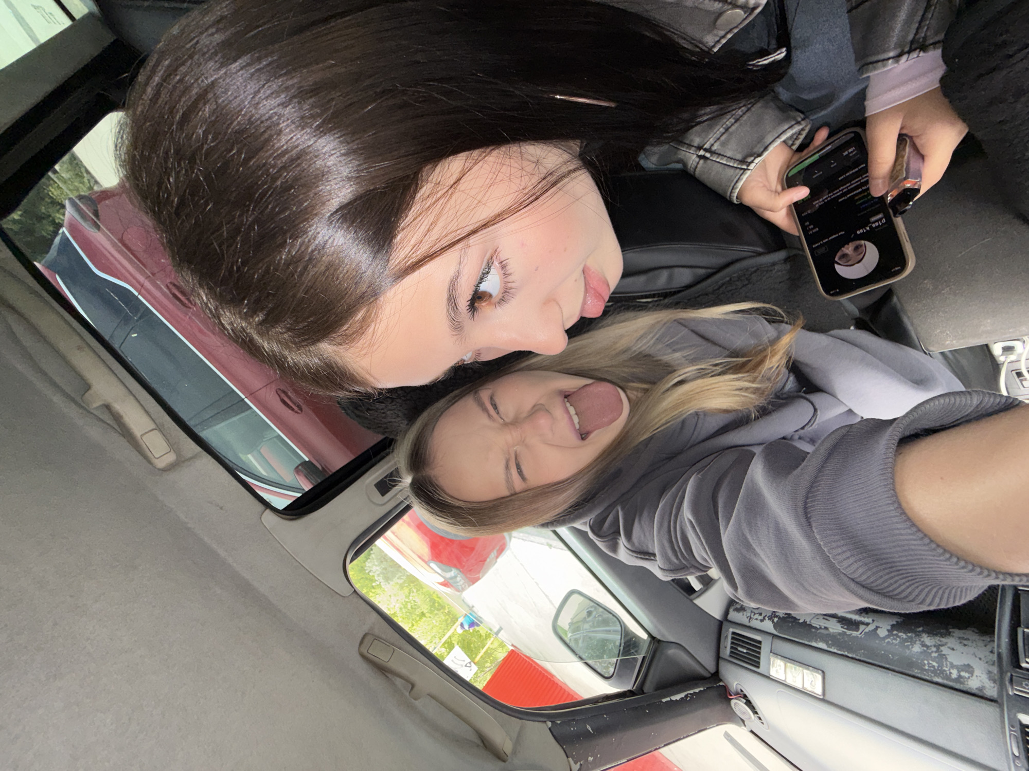 The image captures two young women inside a car, shot from a slightly high and tilted angle, creating a playful, informal snapshot.
The woman in the foreground has long, dark brown hair and is wearing a dark jacket, possibly leather, over a lighter-colored top. She gazes towards her companion with a subtle smile and her tongue slightly extended. In her left hand, she holds a black smartphone, displaying what appears to be a social media screen. The visible text on the screen shows the profile name "pls_kiss" and a profile picture of a person wearing a light head covering. A small, dark device, possibly a vape pen, is attached near the phone.
Behind her, the second woman has blonde or light brown hair and is dressed in a grey hooded sweatshirt. She makes a humorous expression, sticking out her tongue and squinting her eyes playfully.
The setting is the interior of a car, with a light-colored headliner and a dark dashboard, parts of which show signs of wear and peeling. Through the car windows, elements of the outside environment are visible, including portions of other vehicles (one red, one darker) and green foliage, suggesting a daytime scene outdoors. The overall mood is casual and lighthearted, indicative of friends sharing a moment.