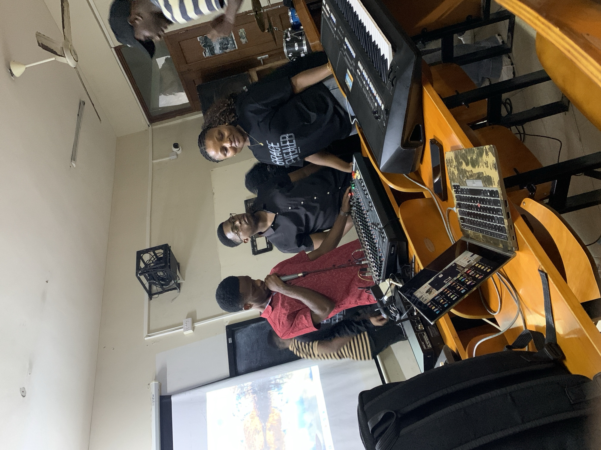 **Scene Description:**
**Location:** Unspecified indoor location in Dar es Salaam, Tanzania.
**Subjects:**
* Four individuals: One wearing a red shirt, holding a microphone, and speaking. Another wearing a black shirt and glasses, operating a sound mixer. A woman wearing a black shirt with the words "Grace Revealed." The fourth person in a striped shirt stands behind them.
**Objects:**
* Musical instruments: Keyboard, drum set.
* Audio equipment: Sound mixer, microphone.
* Technology: Laptop, keyboard, television, projector.
* Furniture: Wooden desks with attached platforms.
**Actions:**
* One individual is actively speaking into a microphone, while another is operating a sound mixer. The woman is playing a keyboard.
**Environment:**
* The setting appears to be a room prepared for a music-related event or recording session.
* A projector is displaying an image on a screen at the front of the room.
* The room has off-white walls and a ceiling fan.
* A security camera is in the room.