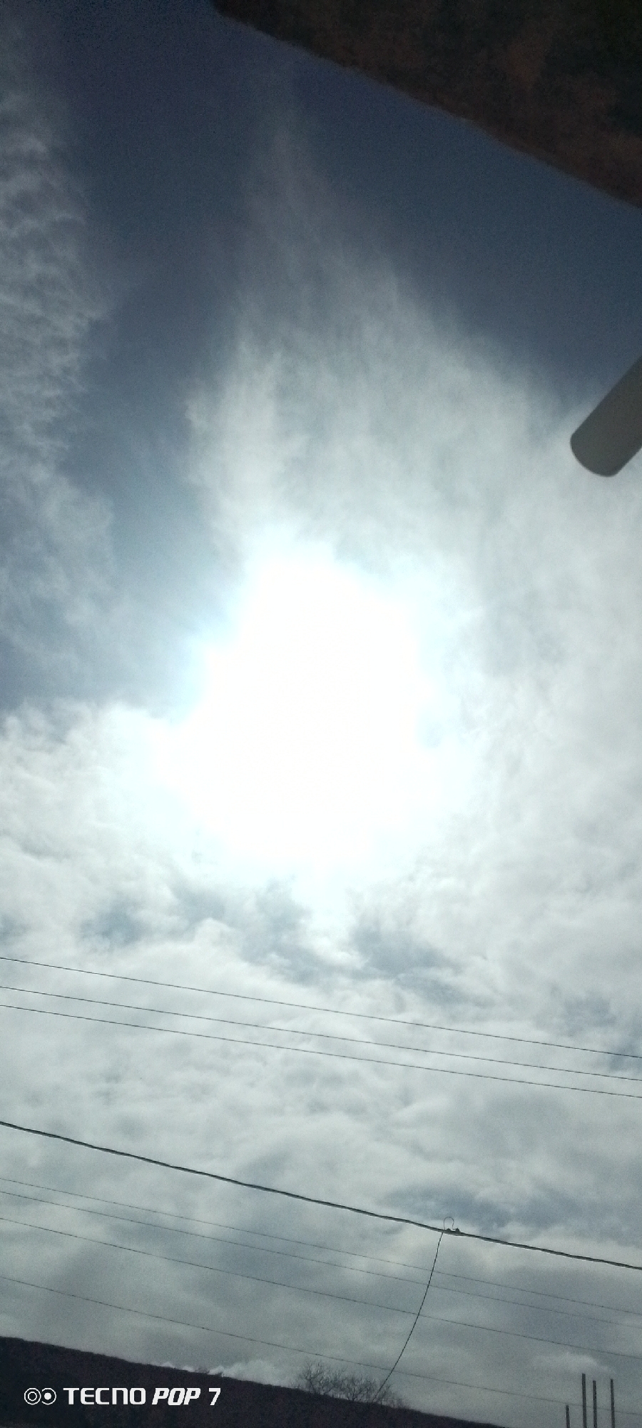 The image is a sky view, possibly taken from the city of Altagracia, Venezuela. The sky is predominantly cloudy, with a bright area suggesting the sun is behind the clouds. Several electrical power lines are visible in the foreground, cutting across the scene. A part of a building or roof is visible at the top of the frame. A small portion of vegetation can be seen at the bottom of the image. The watermark "TECNO POP 7" appears at the bottom left corner.